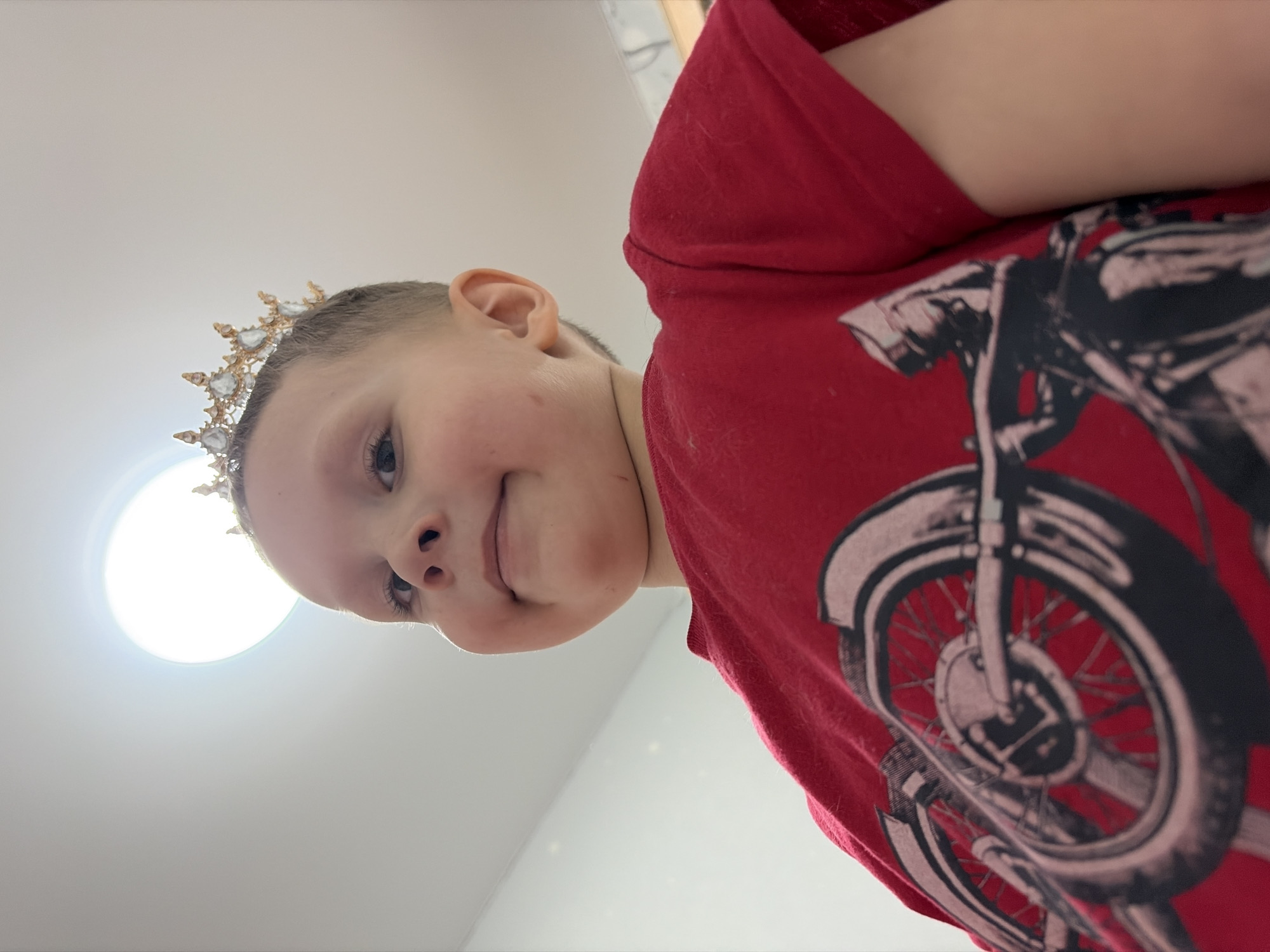 A low-angle shot depicts a young child wearing a jeweled crown. The child has fair skin and is wearing a red t-shirt with a black motorcycle graphic. The background is a white ceiling with a circular light fixture. The location is Unknown, Unknown.