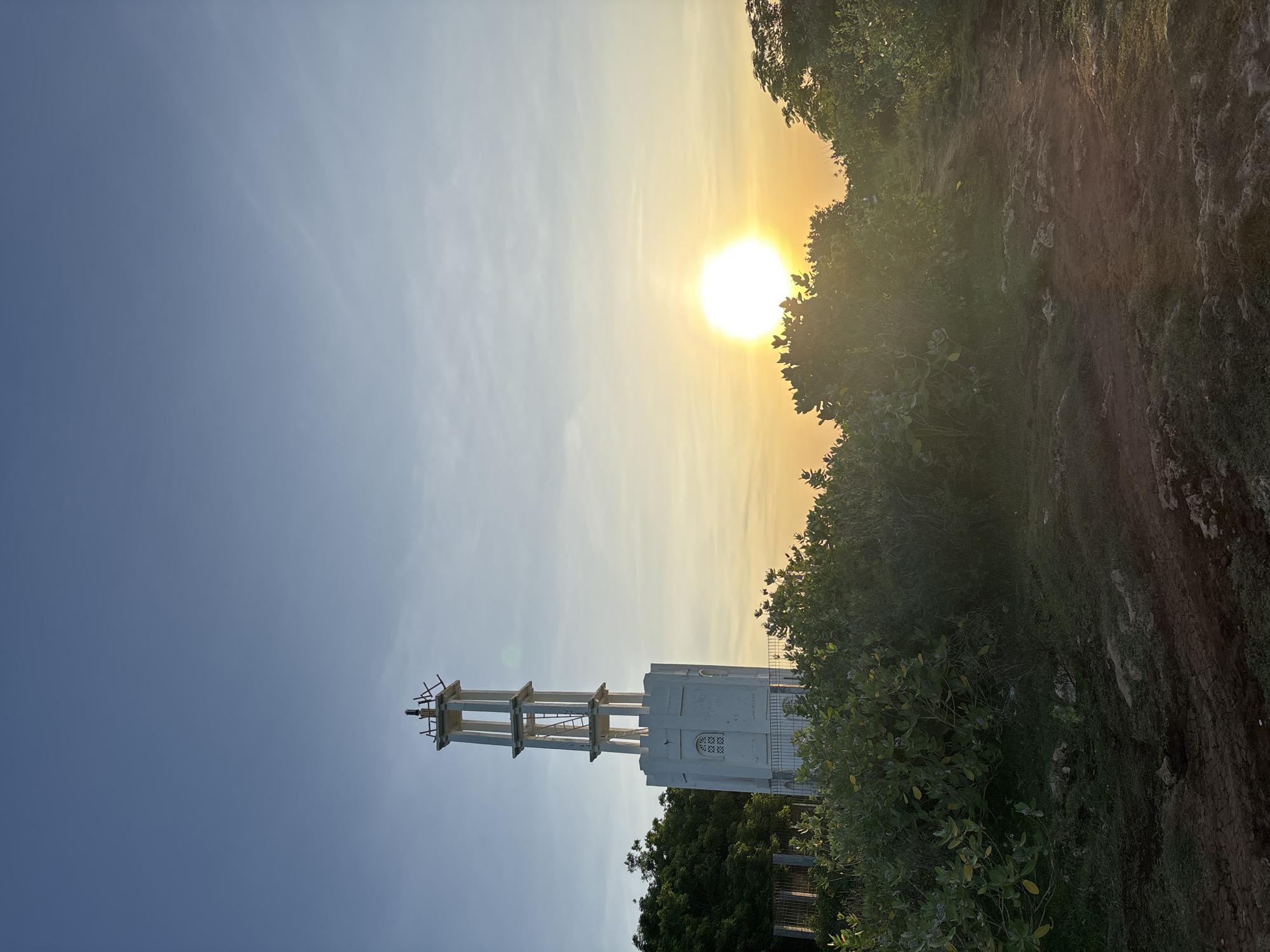 The image shows a vertical view of a white tower-like structure with a ladder attached to its side, set against a backdrop of a bright sky with the sun partially obscured by foliage. The tower's construction is partly exposed, with structural elements visible. The surrounding environment includes dense green vegetation and a rocky terrain, indicating a natural setting. The photograph is taken outdoors during daylight, suggesting a time near sunrise or sunset due to the sun's position. This is the Garuda Wisnu Kencana Cultural Park in Kuta Selatan, Indonesia.