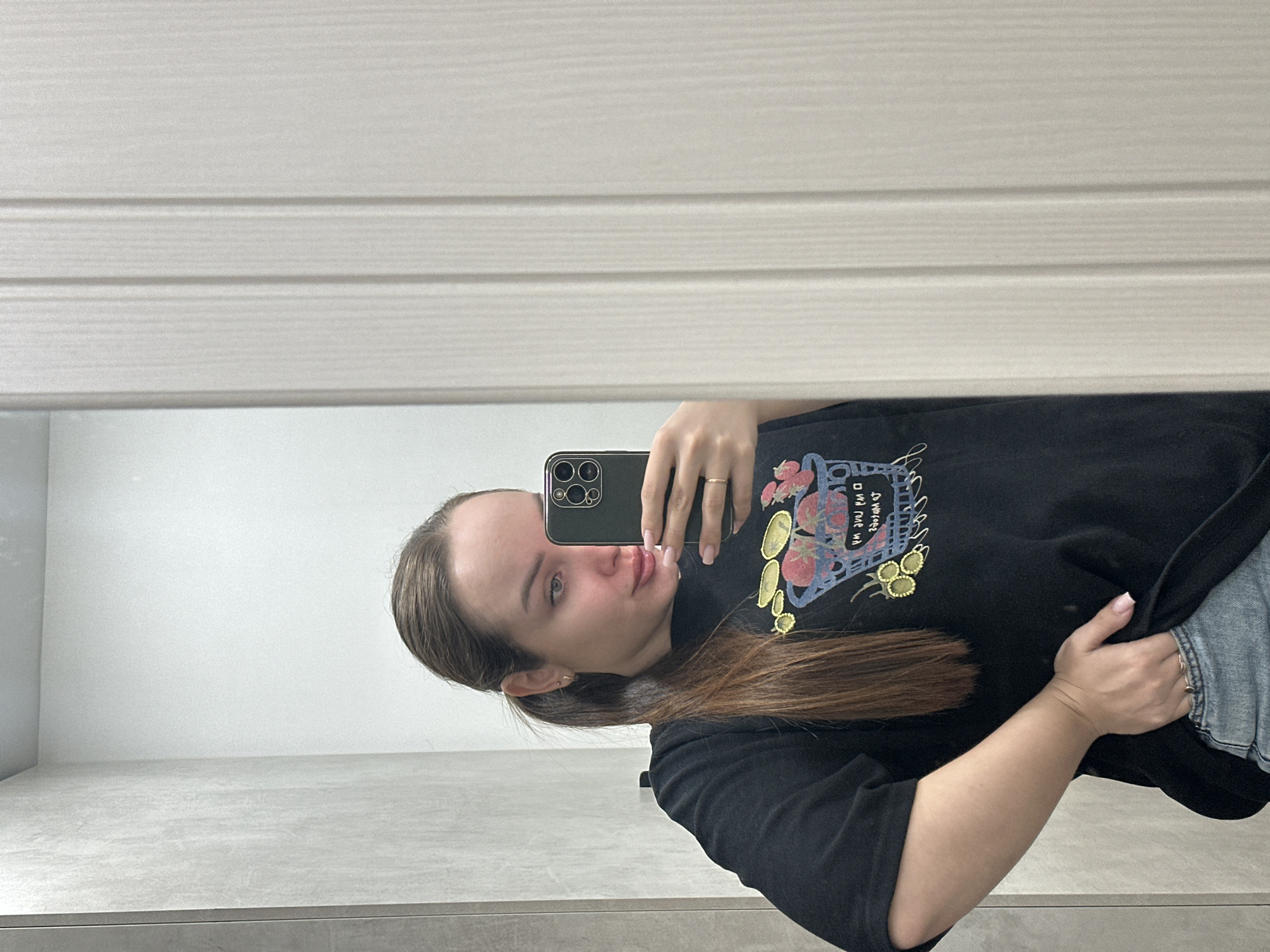 A woman takes a mirror selfie. She is wearing a black t-shirt with a colorful graphic design on the front and blue jeans. She is holding a black smartphone in her right hand. The background consists of a white wall and grey tile. It is impossible to confirm the location in the city of Chaikovsky, Russia from this image alone.