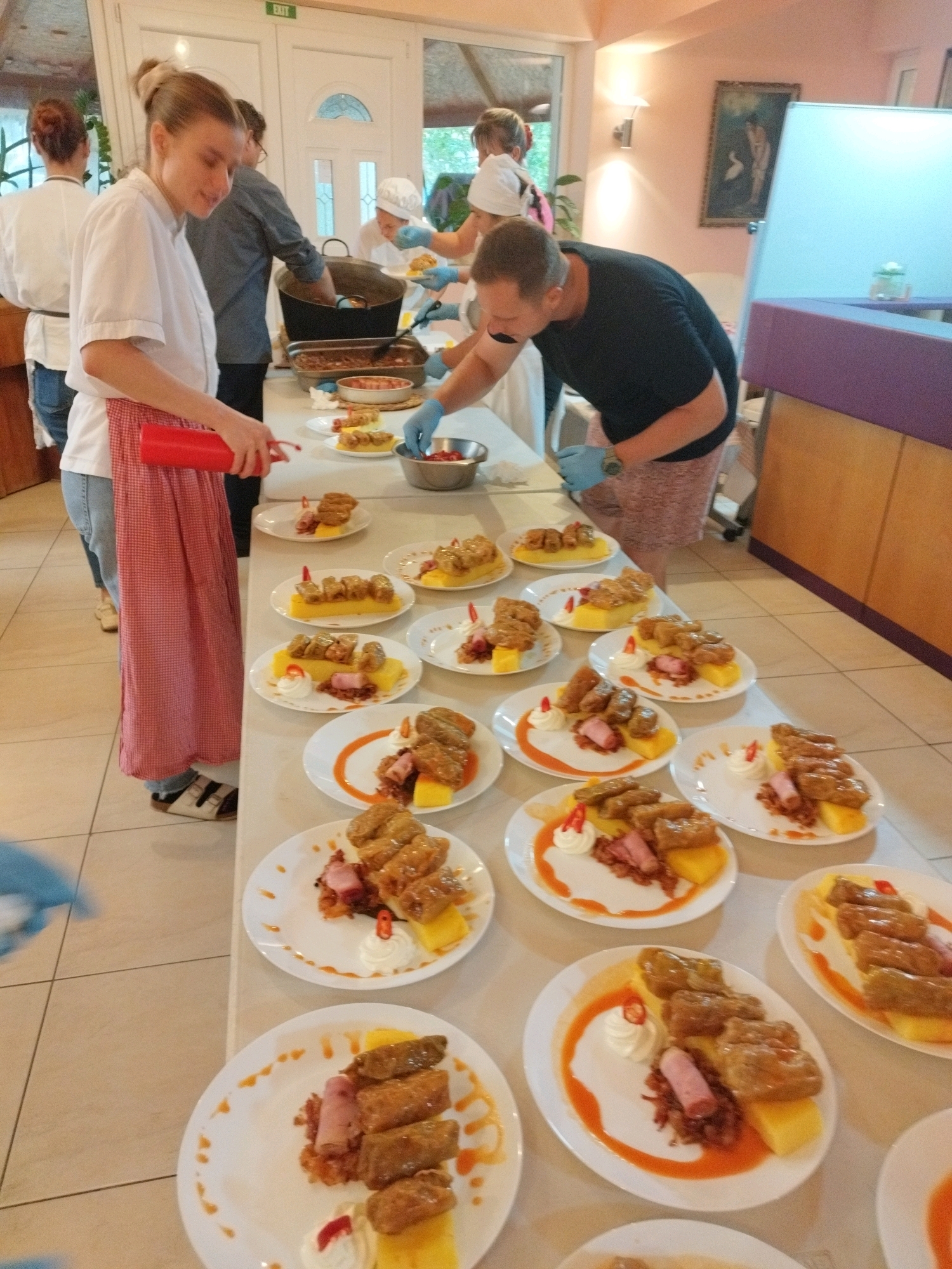 This is an eye-level, full-shot photograph of a buffet-style food service setup, likely a catering event or a restaurant kitchen preparing for service. The scene is well-lit with a warm, ambient glow, suggesting an indoor setting. The predominant colors are the creamy white of the plates and tablecloth, the rich browns and golds of the food, and the pink and red accents in the clothing and garnishes.
In the foreground, a long table is laden with numerous white plates, each holding a portion of what appears to be stuffed cabbage rolls served over a square of polenta, garnished with a dollop of cream and a small red chili pepper. The food is arranged meticulously, indicating a focus on presentation. The plates are staggered and lined up, suggesting a serving line.
To the left, a woman with her hair pulled back in a bun, wearing a white chef's jacket and a red and white checkered apron, is holding a red squeeze bottle, possibly for sauces or garnishes. She is looking down at the plates, engaged in the food preparation. Behind her, another woman in a white chef's hat and uniform is visible, also working.
In the mid-ground, a man in a dark t-shirt and patterned shorts, wearing blue gloves, is leaning over the table, also engaged with the food. He appears to be adding a final touch to the dishes. Further back, several large serving trays are visible, holding more food, and other individuals in kitchen attire can be seen working. One tray appears to contain baked beans.
The environment includes a wall in the background with a framed painting of a nude figure with a swan, and a framed "EXIT" sign above a doorway. The floor is tiled in a beige and cream pattern. A purple upholstered bench or counter is visible on the right.
The overall impression is one of organized culinary activity, with a clear emphasis on preparing and presenting food efficiently and aesthetically. The presence of chef uniforms and gloves suggests a professional food service operation.
Given the detail and style of the food presentation, the ambiance, and the attire of the people, this scene could plausibly be located in a restaurant or catering establishment in Bucharest, Romania, known for its rich culinary traditions and vibrant food scene.