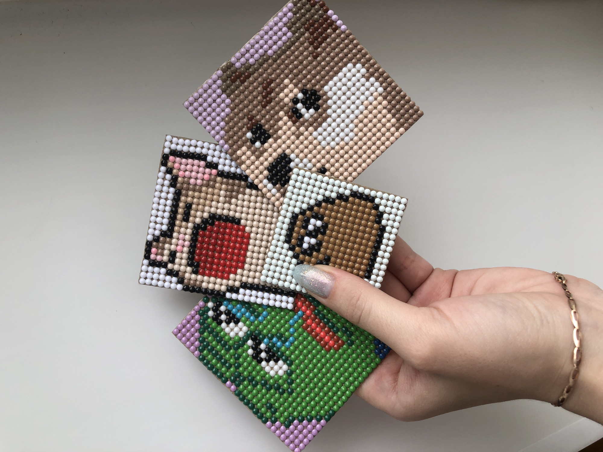 A close-up shot depicts a hand holding a stack of four small, square canvases decorated with a mosaic of tiny, colorful beads. The mosaics depict cartoonish images, including a dog face, possibly another animal, and a green image with a red element. The hand has a single glittery nail. A gold bracelet is visible on the wrist. The background is a plain white surface, likely a tabletop or counter. The location, Noviy, Russia, is not discernible from the image content.