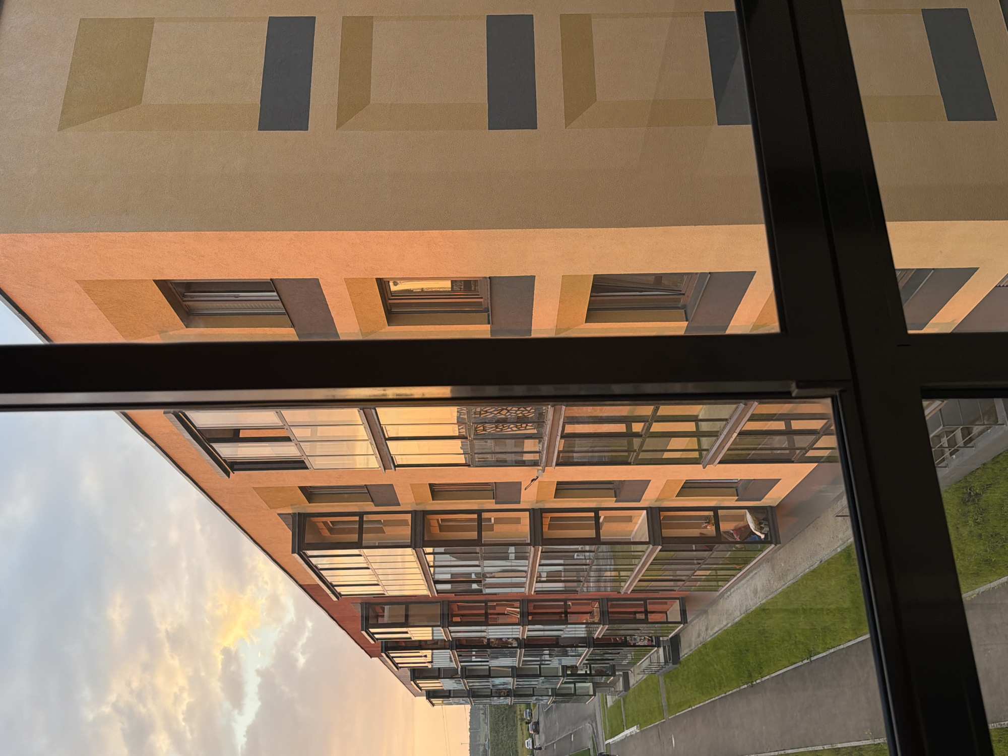 The image captures a modern apartment building in the city of Froly, Russia, during sunset. The building features a warm, orange-toned facade with geometric decorative elements. Many of the apartments have glass-enclosed balconies, reflecting the golden hues of the sky. A paved walkway and a strip of green grass run along the side of the building, with a few cars visible on a distant road. The sky is filled with soft, warm clouds, indicating the end of the day. The photo is taken from inside another apartment, with a dark window frame partially obscuring the view of the building.