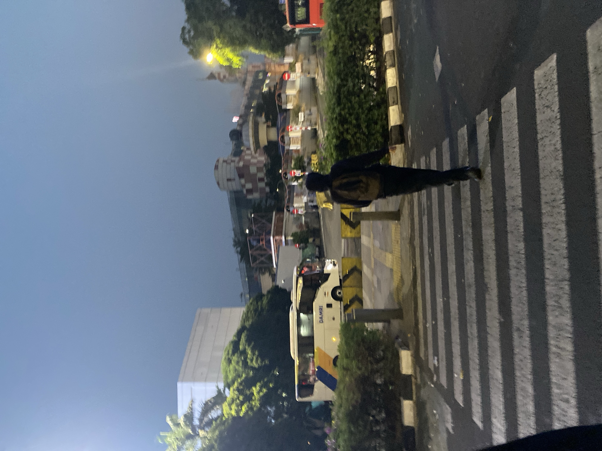 A person wearing a backpack walks across a pedestrian crossing in front of a white bus with blue and yellow stripes that has "DAMRI" written on its side. In the background, there is a multi-story building with red and white checkered patterns, trees, and streetlights. The scene appears to be at dusk or night in a city. This is likely the area around FX Sudirman Mall, a prominent landmark in South Jakarta, Indonesia, known for its distinctive architecture and proximity to transportation hubs like the TransJakarta bus stops.