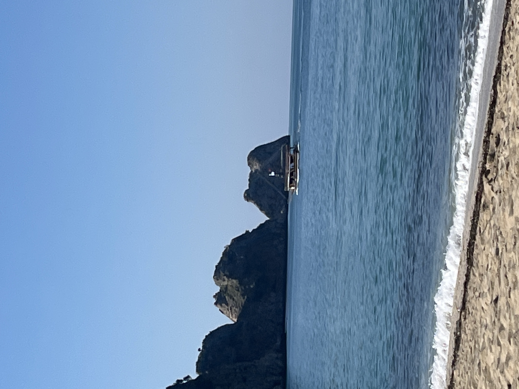The image depicts a coastal scene, possibly near Alanya, Turkey. To the left, a rocky formation rises from the water. A small boat is sailing alongside the rock formation. The ocean is blue and stretches towards the horizon. A sandy beach is on the right. The sky is clear and blue.