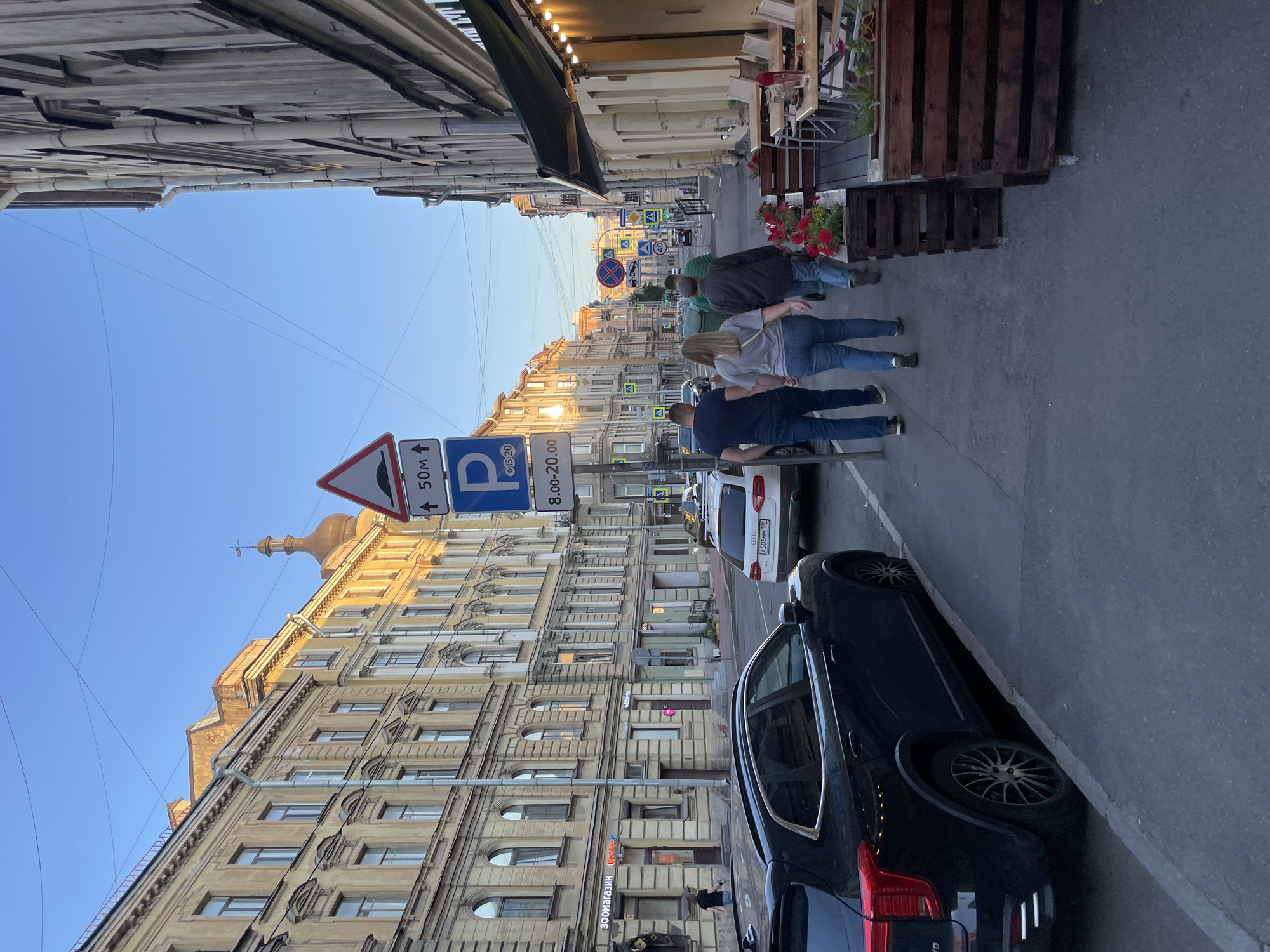 This image depicts a street scene in Saint Petersburg, Russia, observed during golden hour, likely late afternoon or early evening, characterized by bright, clear blue skies and long shadows. The sun casts a warm, golden light on the upper facades of the buildings on the left side of the street, contrasting with shaded areas.
The street, paved with dark asphalt, runs diagonally into the distance. On the left side, a black multi-purpose vehicle (MPV) with dark alloy wheels is parked parallel to the curb, its rear illuminated by the sun's reflection. Further down, a white Audi sedan (license plate A505MM 98) is also parked. The sidewalk on the left features various storefronts with Cyrillic signage, including "ЗООМАГАЗИН" (Pet Shop) and "СтройБург" (Construction/Building). The buildings on this side are multi-story, exhibiting classical architectural elements such as ornate window frames, cornices, and decorative relief work, rendered in pale yellow and cream tones. In the mid-ground, a distinctive golden dome with a spire and weather vane is visible above the rooftops.
In the center-right foreground, four individuals are walking away from the viewer on the right-hand sidewalk. From left to right, the closest is a man in a dark blue t-shirt and jeans, followed by a woman with light brown hair in a light grey t-shirt and blue jeans, then a man in a dark green t-shirt and jeans, and a fourth person further back in a striped t-shirt.
Adjacent to the walking pedestrians, on the right side of the street, an outdoor dining area is established, featuring several light-colored tables and chairs. Dark wooden planters containing red flowering plants line the edge of this area. An awning, dark in color, extends over the seating, adorned with a string of warm-toned lights. The building on the right also appears to be multi-story with a muted, possibly grey or beige, facade and prominent vertical drainpipes.
A prominent cluster of Russian road signs stands near the center of the image. These include a triangular warning sign for a speed bump ("лежачий полицейский") with an "↑ 50m" indicator, and a blue rectangular sign denoting paid parking ("P" with coin symbol) with operating hours "8.00-20.00". Further down the street, additional signs indicate "No Entry," "No Stopping," a pedestrian crossing, and a "40" speed limit. Overhead, numerous utility wires crisscross the sky.