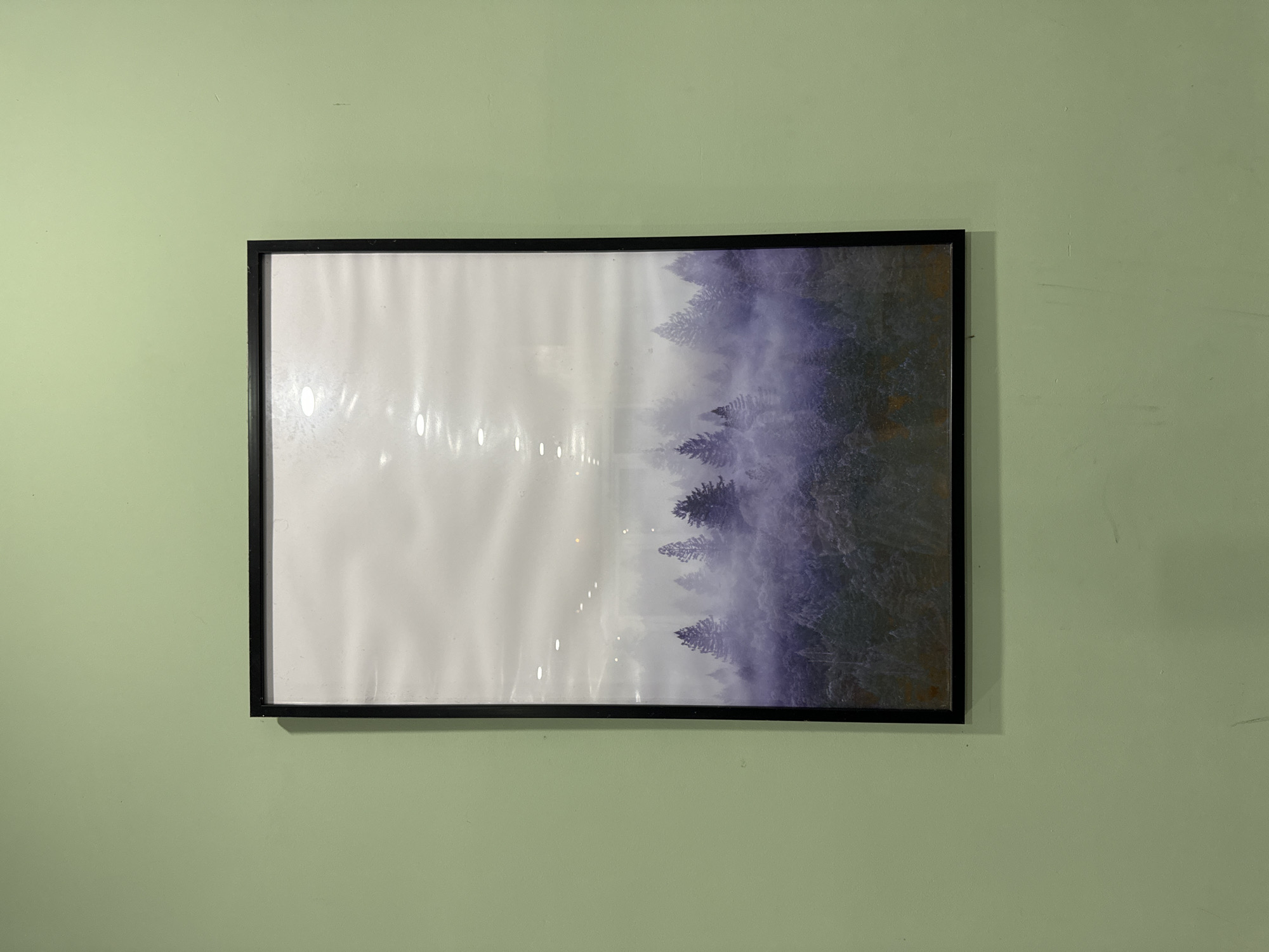 A framed print of a forest scene hangs on a light green wall. The print is in a black frame and depicts a forest shrouded in mist or fog. The left side of the print is lighter, gradually darkening towards the right where the forest is most visible. The print has reflections. No people are visible. No information in this image indicates this location to be Da Lat, Vietnam.