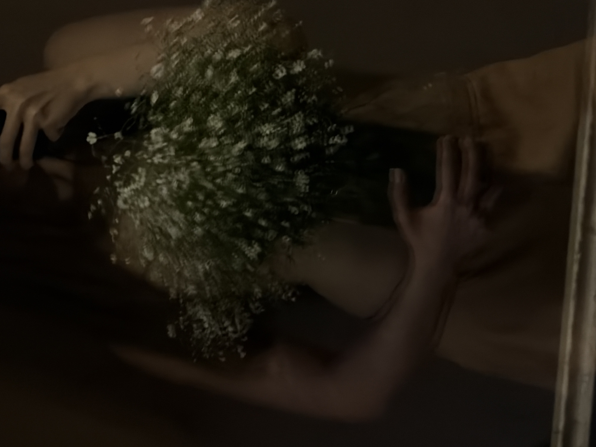 A person is holding a bouquet of small white flowers in a dark room. The person's face is not visible, but they appear to be taking a selfie in a mirror. The room is dimly lit and appears to be unfurnished. The person is wearing a tan-colored garment.