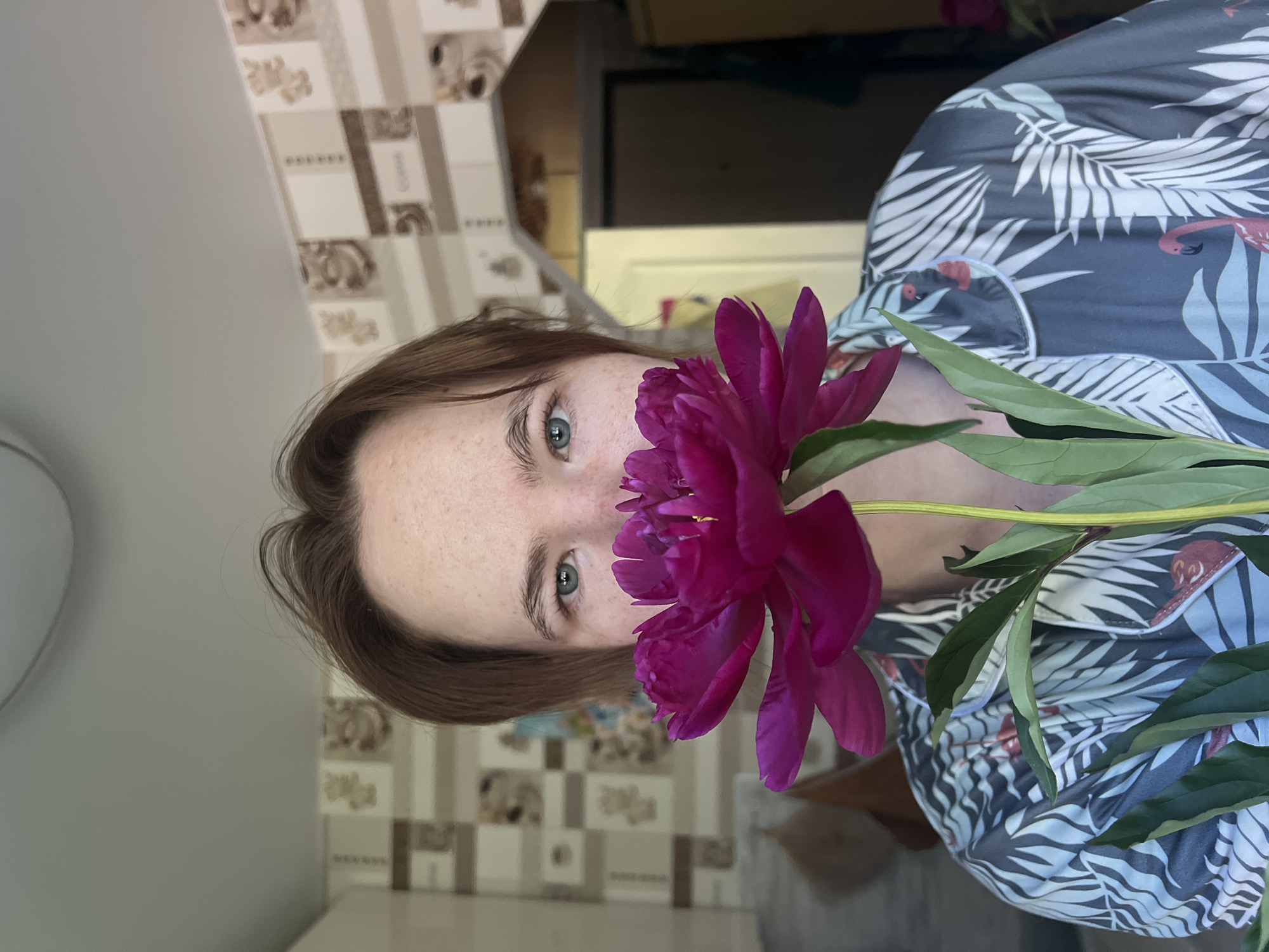 The image shows a person holding a deep pink peony flower in front of their face, obscuring their mouth and nose. The person has light skin with freckles and light blue eyes. They have short, brown hair and are wearing a dark gray button-up shirt with a tropical leaf and flamingo pattern. The background appears to be an interior space with patterned wallpaper and a doorway in the background. The image was captured in Chaikovsky, Russia.