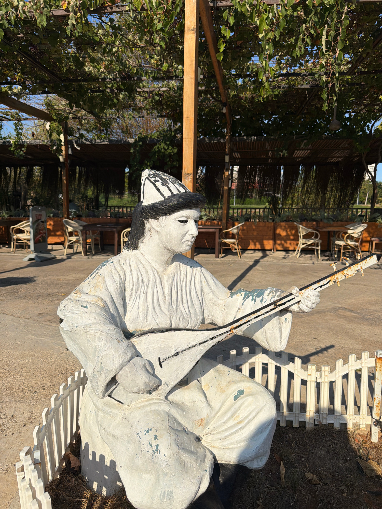 The image depicts a statue of a musician playing a traditional stringed instrument, possibly a dutar or tanbur, in an outdoor setting. The statue is white with black accents on the hat and hair, and it appears to be made of a textured material, showing signs of wear and chipping paint. The musician is seated and dressed in loose, flowing robes, characteristic of Central Asian attire. A small white picket fence surrounds the base of the statue, which is partially covered with dried grass and leaves.
In the background, there is a shaded outdoor seating area with wooden tables and chairs, covered by a canopy of green vines. This area appears to be part of a restaurant or cafe. The flooring is paved with light-colored gravel or sand. Beyond the seating area, there is a glimpse of greenery and possibly other structures, suggesting a park-like or recreational environment. The overall lighting suggests it is daytime, with clear sunlight casting shadows. While the exact location is not visible in the image, the presence of grapevines covering an overhead structure is a common feature in outdoor dining areas, and this style of architecture is found in various tourist areas. Given that the image was taken in Antalya, Turkey, this scene could be from a garden restaurant, a historical park, or a cultural center within the city.