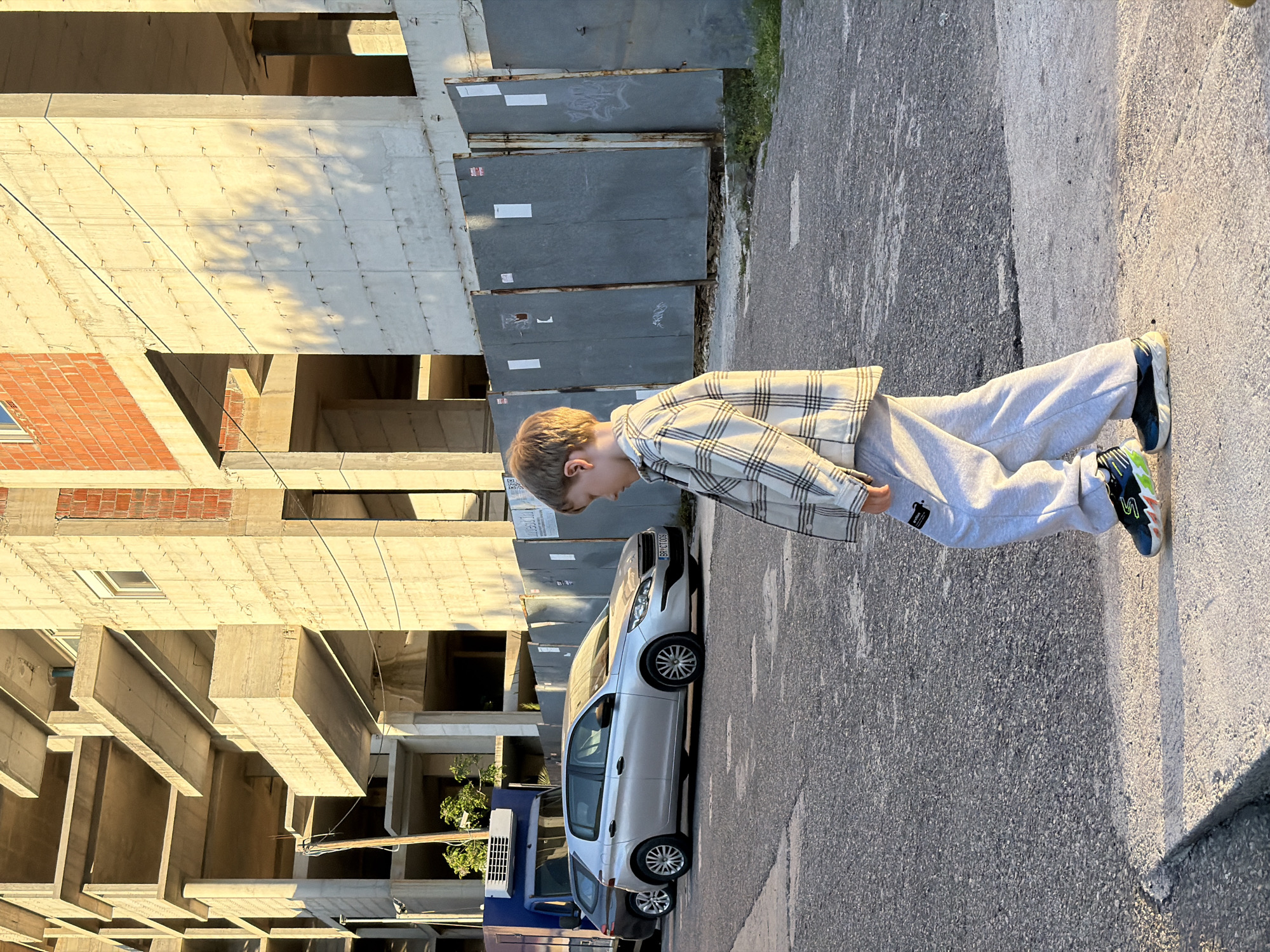 A young boy stands on an asphalt road in Petrovac centar, Montenegro. He is wearing a plaid shirt and gray sweatpants. Behind him is a silver car parked in front of a building under construction. The building is made of concrete and brick. There are also some gray panels or sheets propped up against the building. A blue truck is partially visible behind the silver car.