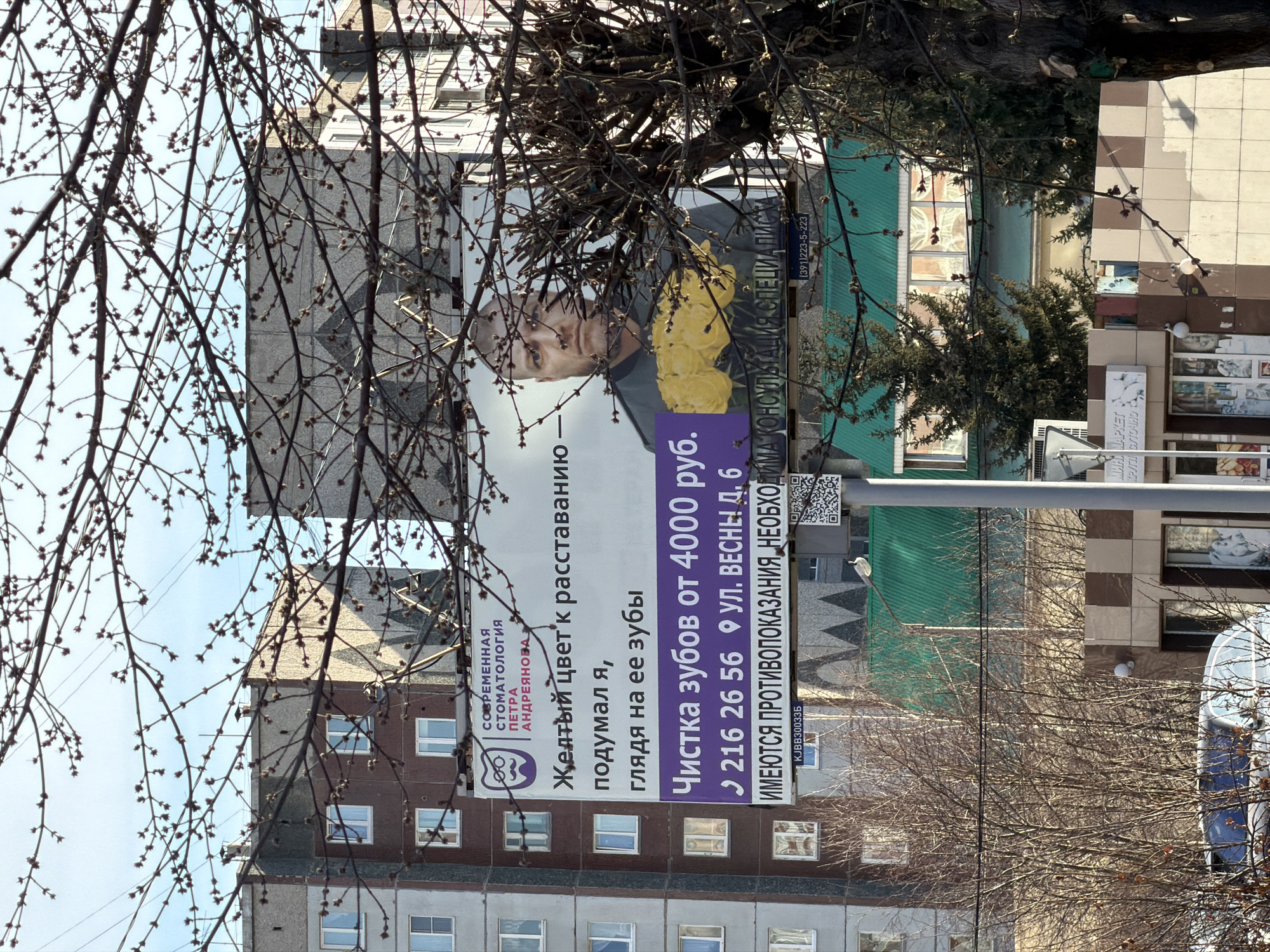 Here is a description of the image:
The image shows a billboard ad for a dental clinic in Krasnoyarsk, Russia. The ad features a man's face next to yellow flowers, and promotes tooth cleaning services for 4000 rubles. The text also mentions the address: Vesny Street, building 6, and a phone number. The sign says, “Modern Dentistry named after Peter Andrianov” . In the background, there are buildings, trees without leaves, and other elements of a city street.