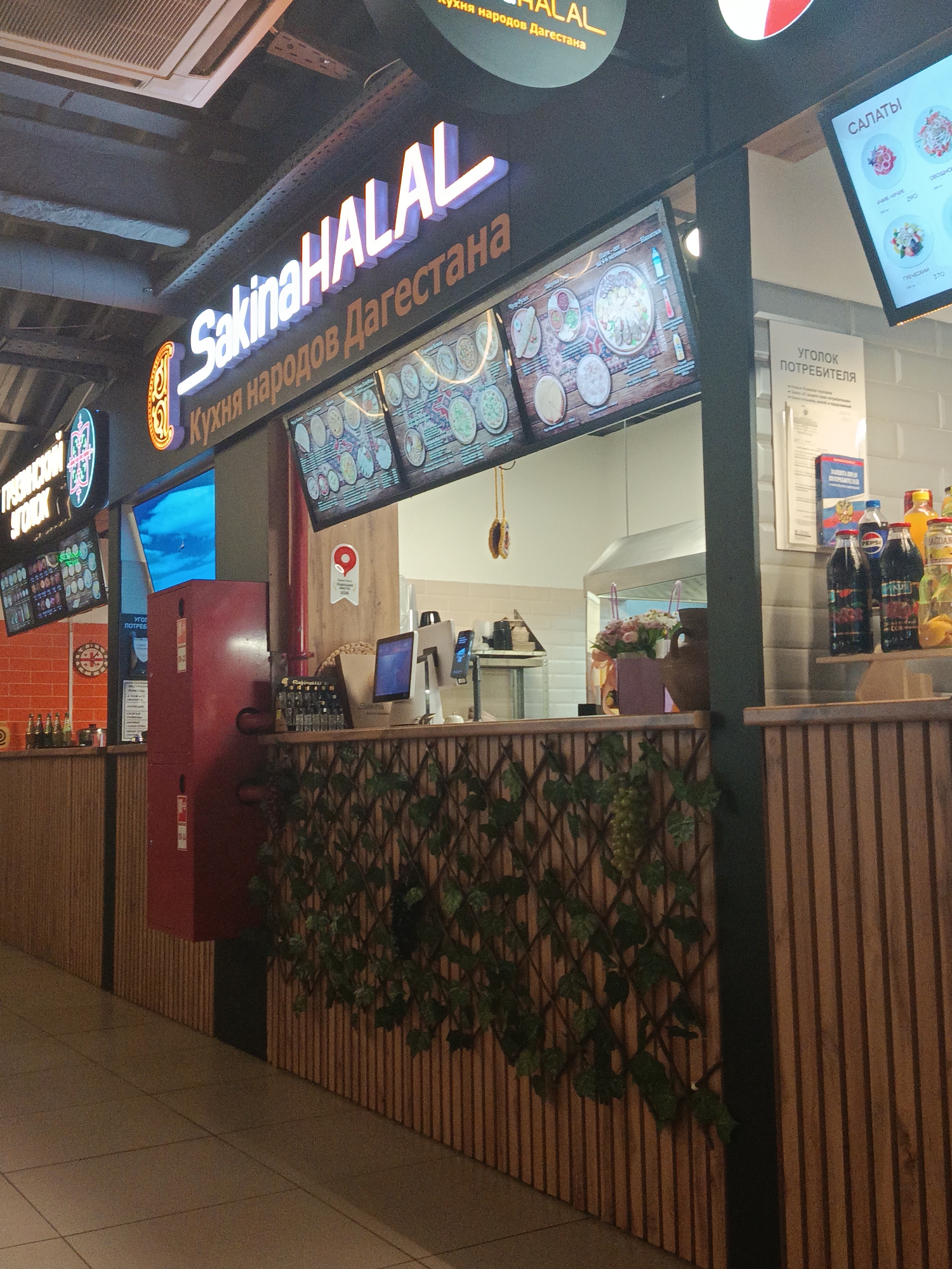 The image depicts the interior of a food establishment with a modern, clean aesthetic. A prominent illuminated sign reads "Sakina HALAL" in large white lettering, with smaller Russian text below it indicating "Кухня народов Дагестана" (Cuisine of the peoples of Dagestan). To the right, a digital menu board displays food items under the heading "САЛАТЫ" (Salads). Another menu board to the left shows various dishes, including what appears to be pizzas and other food items.
The establishment features wooden paneling on the walls and counters, creating a warm and inviting atmosphere. A red fire extinguisher is mounted on the wall. Several bottles of drinks, including Pepsi, are visible on a shelf behind the counter. A small bouquet of flowers in a vase adds a decorative touch. In the background, a stainless steel exhaust hood is visible, suggesting a kitchen area. The floor is tiled. The overall scene is well-lit, indicating an indoor commercial space.