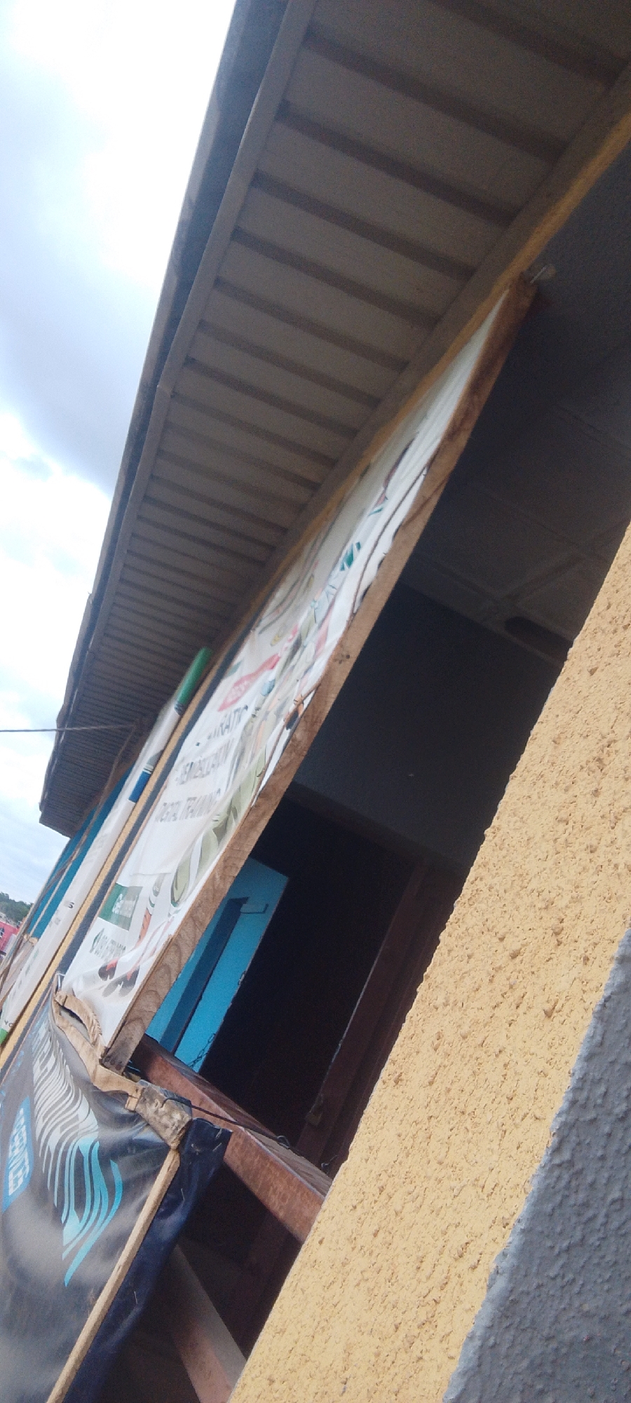 The image shows the exterior of a shop with a corrugated iron roof extending outwards. Below the roof, a white banner with various graphics and text is partially visible, attached to a wooden frame. The banner appears to be advertising services related to "Digital Training." To the right of the banner, a dark opening suggests an entrance or window into the shop, with a glimpse of a blue interior. The shop's exterior wall is textured and painted in a yellowish hue. The overall scene depicts a simple storefront in what appears to be an outdoor setting, possibly during daytime. The lighting suggests it's not midday, as shadows are not prominently cast. The sky is visible in the upper left quadrant, appearing overcast. No people are directly visible in the frame. The banner has text such as "EMBELLISHIN" and "DIGITAL TRAINING". There are also logos or icons on the banner, including one that resembles a green leaf. The image is taken from a low angle, looking upwards towards the roof and banner.