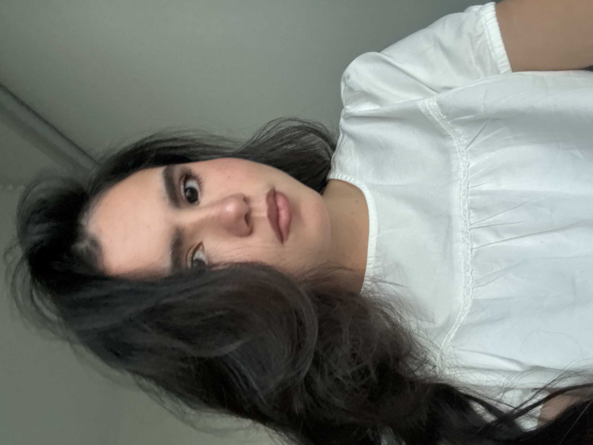 A close-up, shoulder-up portrait of a young woman with dark, voluminous hair frames the left side of the image. She is looking directly at the camera with a neutral expression. Her skin is fair with a scattering of freckles across her nose and cheeks. She is wearing a white, short-sleeved top with a ruffled neckline and delicate lace trim. The background is a plain, light gray wall, indicating an indoor setting. The lighting is soft and natural, suggesting it may be daytime. There are no visible landmarks or context to determine a specific location within the city of Austin, United States of America.