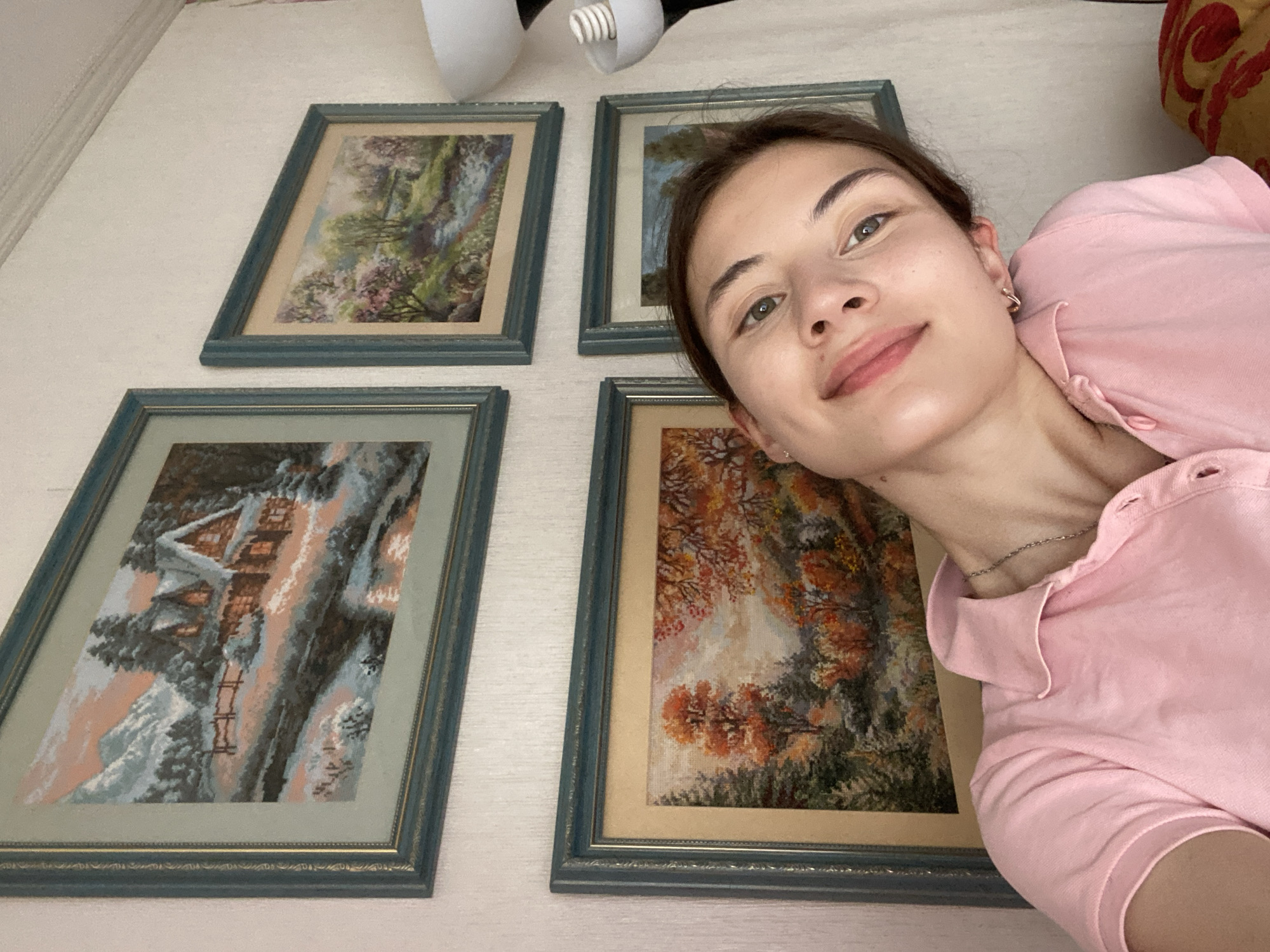 The image shows a young woman with fair skin, green eyes, and brown hair, wearing a pink polo shirt and a silver necklace. She is leaning her head to the side, smiling at the camera. Behind her, on a textured white wall, are four framed pictures depicting nature scenes. The top left picture is a winter landscape with a house. The top right and bottom right pictures appear to be spring and summer scenes with trees and water. The bottom left picture is an autumn landscape with colorful trees. The lighting suggests an indoor setting, likely a home. The presence of framed artwork on the wall implies a domestic environment. There is no visible indication of the specific location within Votkinsk, Russia.