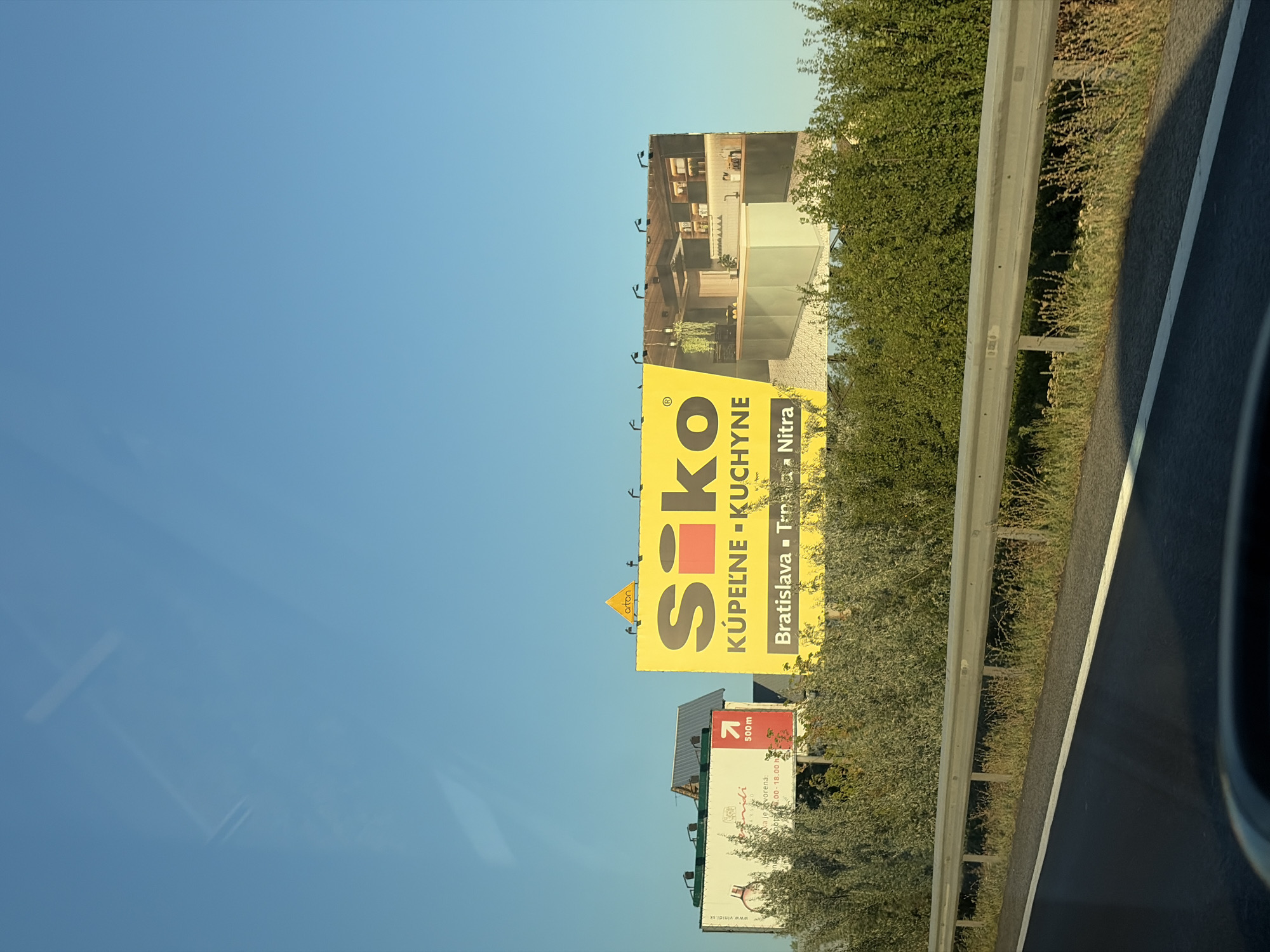 This is a photograph taken from a moving vehicle on a clear, sunny day, showing a roadside billboard in Báb, Slovakia. The billboard is yellow and features the text "Siko" in large black letters, with the words "KÚPELNE - KUCHYNE" (bathrooms - kitchens) below it. To the right of the text is an image of a modern kitchen. Below "KÚPELNE - KUCHYNE" are the words "Bratislava - Trnava - Nitra". There is another, smaller white billboard to the left, which appears to advertise a company called "Vindi" and shows a perfume bottle. The billboards are situated behind a metal guardrail and surrounded by green bushes and trees. The sky is a clear, pale blue, with some reflections visible from the vehicle's windshield.