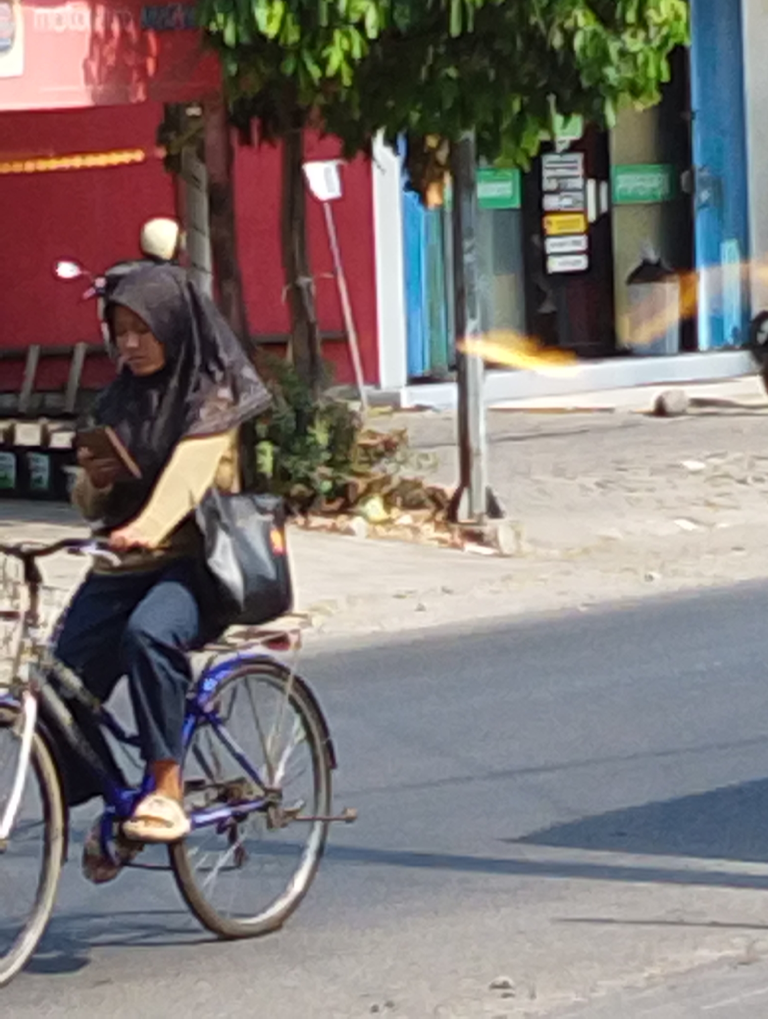 A woman wearing a black hijab and a brown top is riding a bicycle down a street in Manaruwi, Indonesia. She is holding a smartphone in her left hand and looking down at it. Her right hand is resting on the handlebars. She is wearing dark blue pants and light-colored sandals. A black bag is strapped to the back of her bicycle. The bicycle has blue accents on the frame and wheels. In the background, there is a red building with "moto" written on it, green trees, and a light blue building with signs on it. The street is paved and appears to be in a commercial area. The lighting suggests it is daytime.