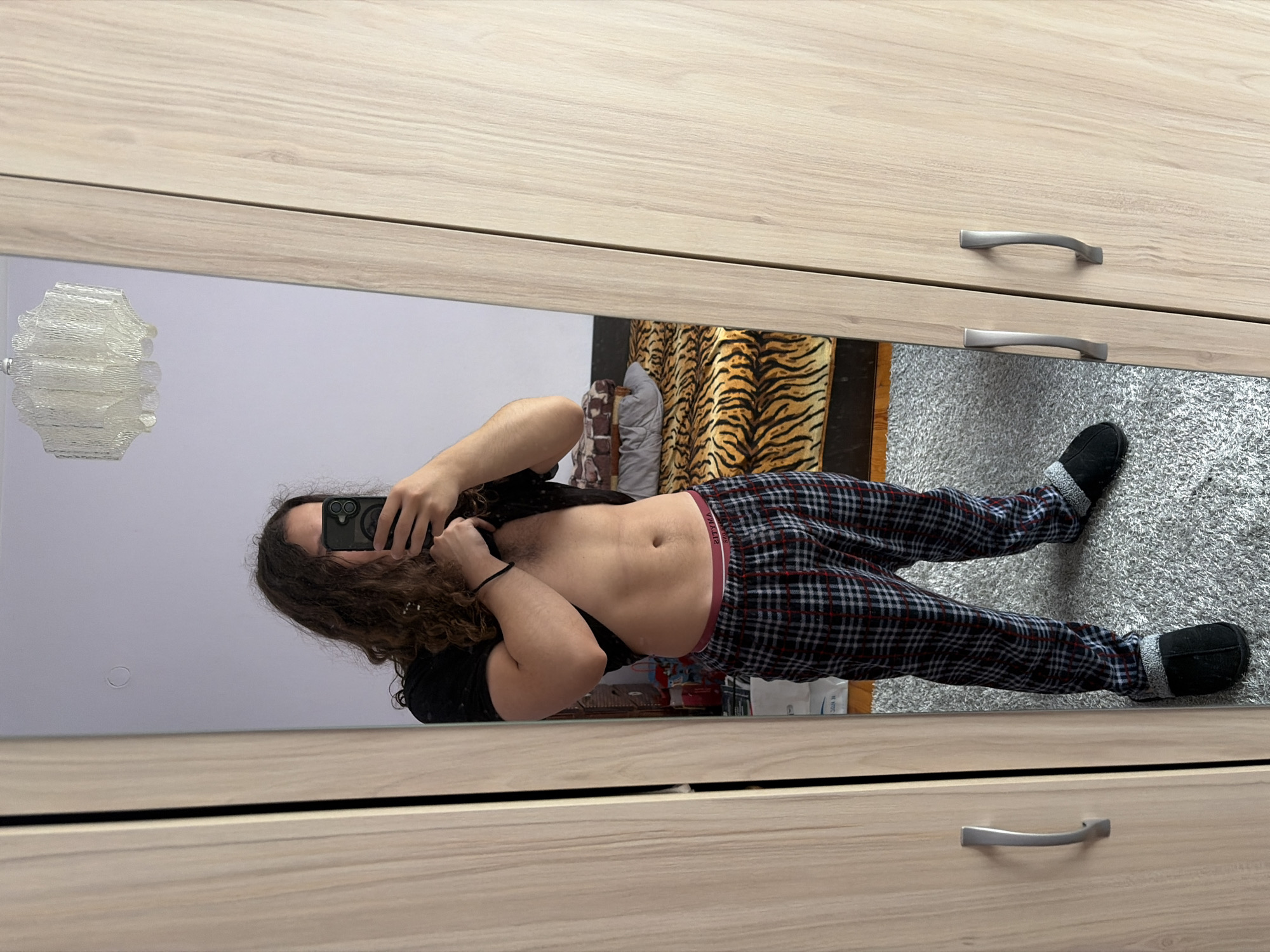 A full-length reflection of a person is visible in a mirror set into a light-colored wooden dresser. The person is wearing plaid pajama pants, black slippers, and a black t-shirt. Their midsection is exposed. They are holding a smartphone and taking a selfie. The background includes a white wall, a decorative wall lamp, a silver rug, and a tiger-striped decorative item. The location cannot be determined from the image.
