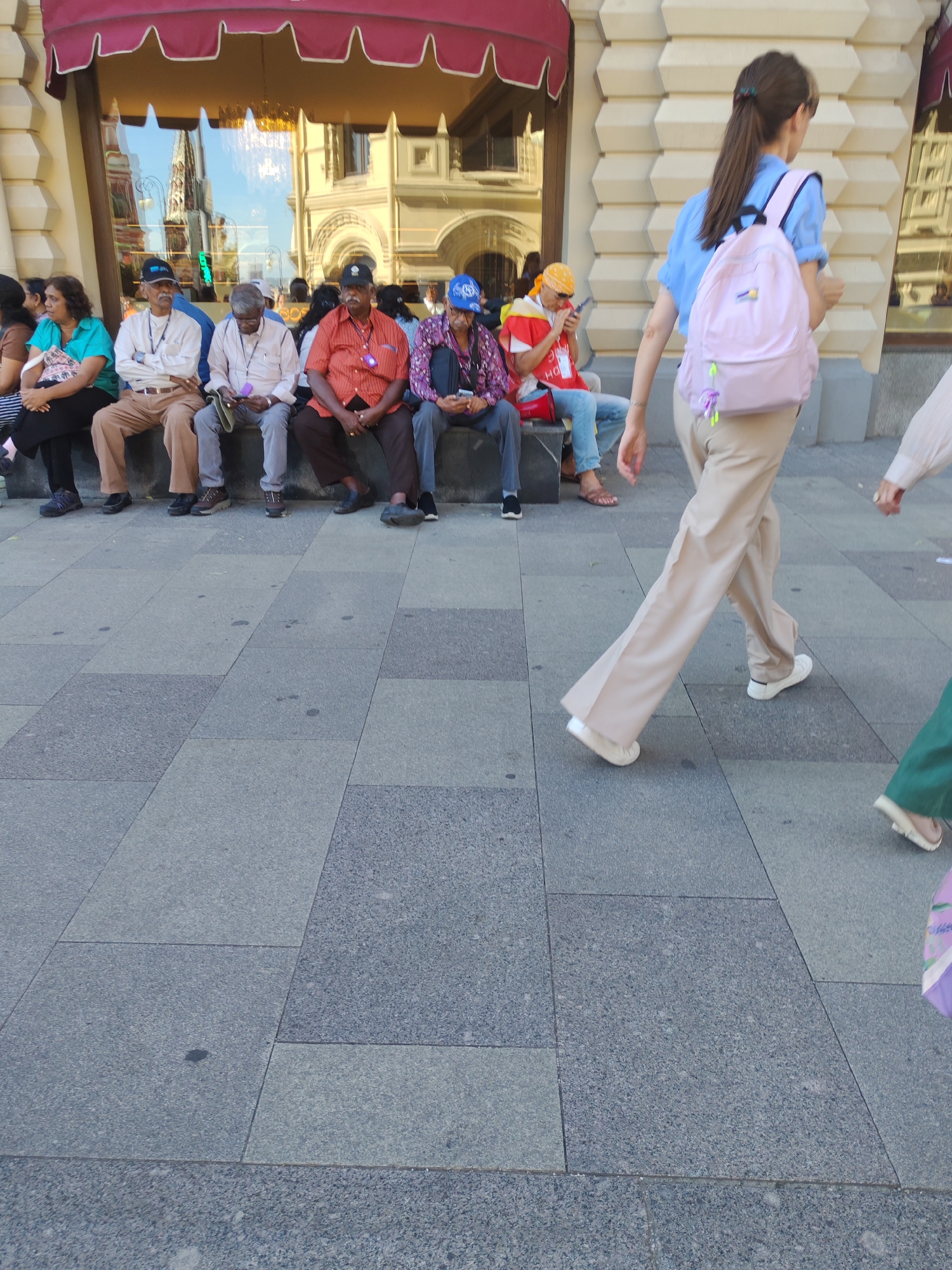 The image shows a group of people sitting on a bench outside a building. The people are of various ages and ethnicities. They are all dressed casually. The building in the background appears to be a commercial establishment with large windows. The ground in front of the bench is paved with stone tiles. A young woman with a pink backpack is walking past the group on the right side of the image. The scene appears to be taking place on a sunny day.