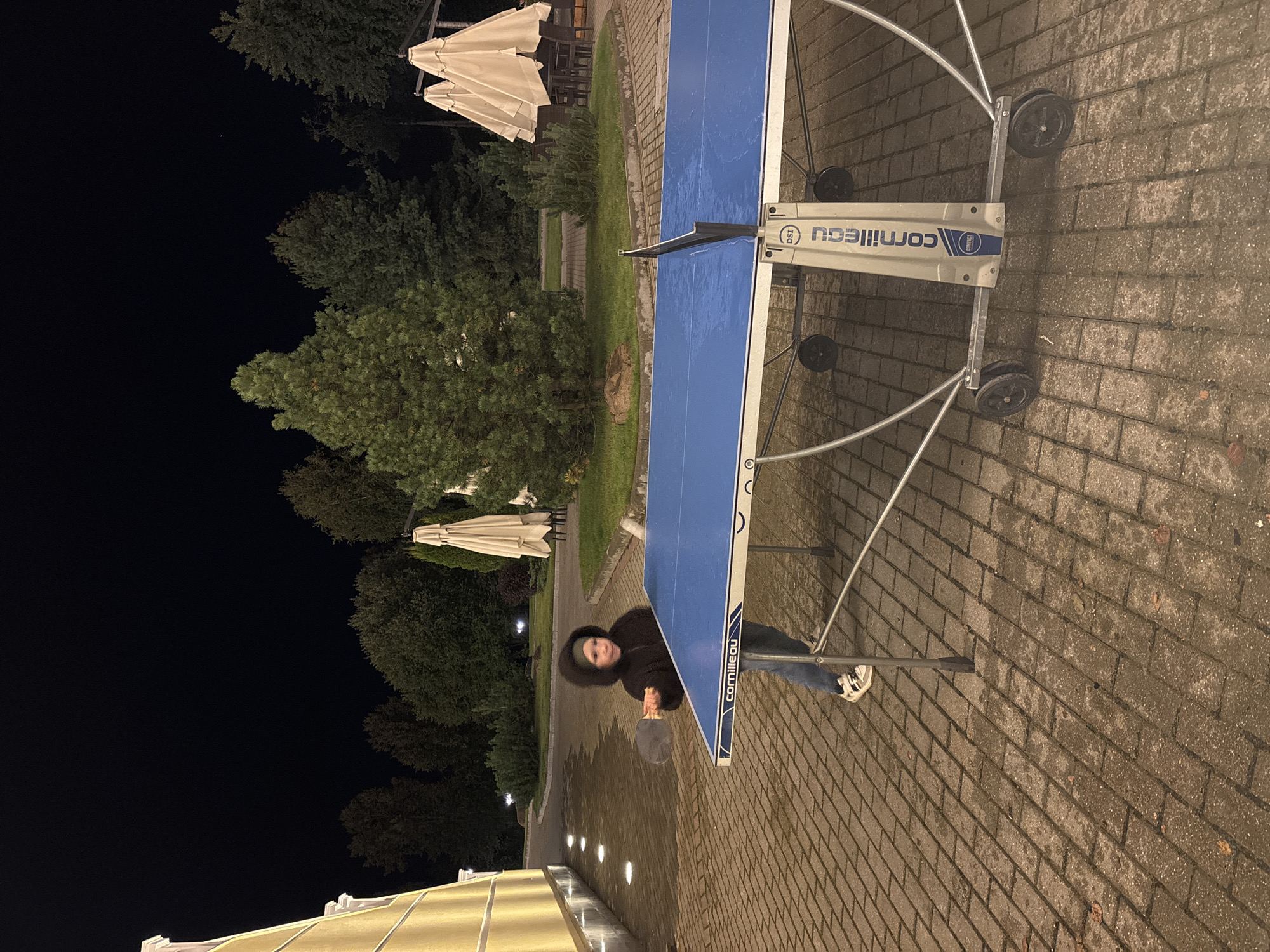 An image taken at night shows a young person playing ping pong outdoors on a paved area in Рождествено, Russia. The person, wearing a dark hooded sweatshirt and jeans, is holding a ping pong paddle and appears to be mid-game, facing the blue ping pong table. The table is wet, suggesting recent rain or high humidity. The surrounding environment includes trees, some with lights illuminating them, and a building with yellow siding on the left. Several closed, light-colored patio umbrellas are visible in the background, along with tables and chairs. The ground is a cobblestone-like paving. The overall scene suggests an evening recreational activity in a semi-public outdoor space.