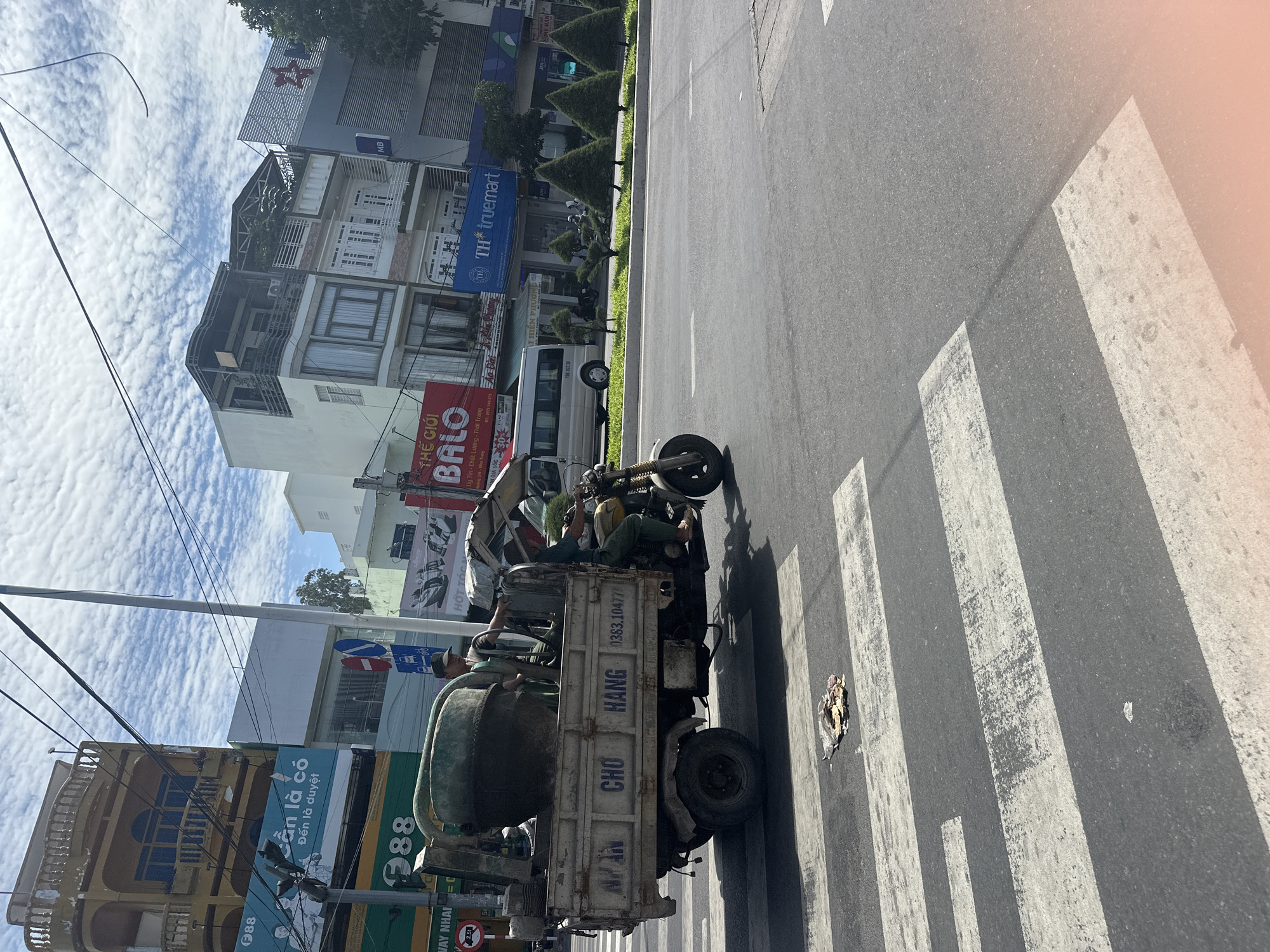 A three-wheeled motorcycle is crossing a pedestrian crossing in Bac Nha Trang Ward, Vietnam. The motorcycle is carrying a large cement mixer. Two men are on the motorcycle; one is driving and the other is standing in the back, holding onto the cement mixer. The motorcycle is yellow and has a white canopy. The cement mixer is green. The pedestrian crossing is black and white. In the background, there are several buildings and trees. The sky is blue with white clouds. A trash pile is on the pedestrian crossing.