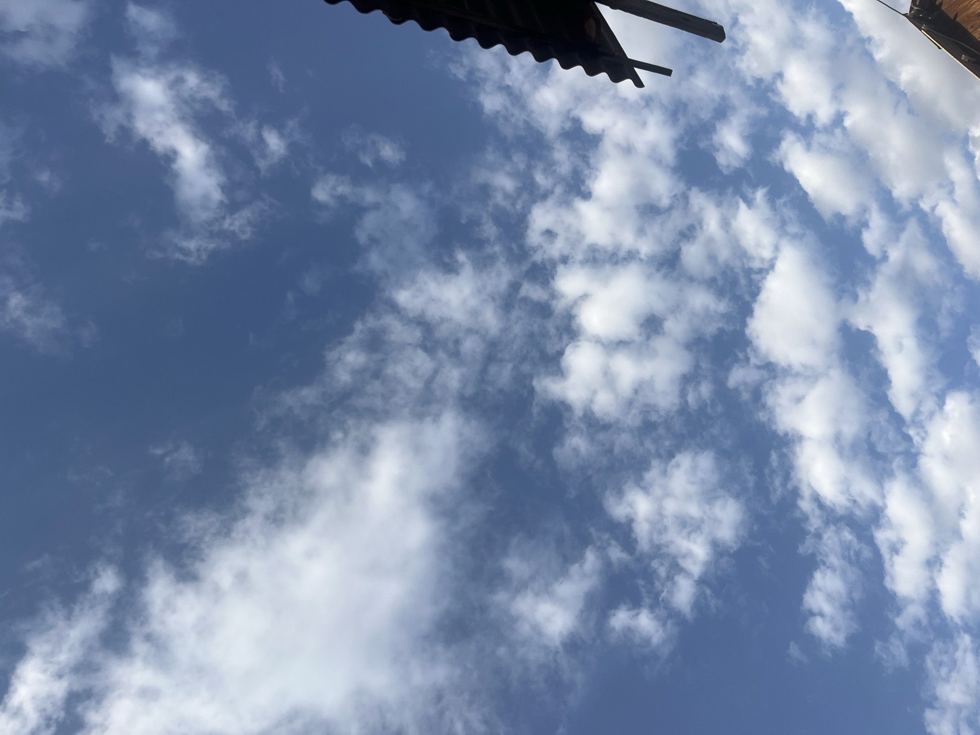 The image shows a bright blue sky with scattered white clouds. In the upper right corner, a portion of a dark, corrugated roof with visible wooden supports is visible, suggesting an outdoor setting. The perspective is looking upwards from ground level. There are no people or animals in the image. The scene depicts a clear day with fair-weather clouds. The location is identified as Batken City, Kyrgyzstan.