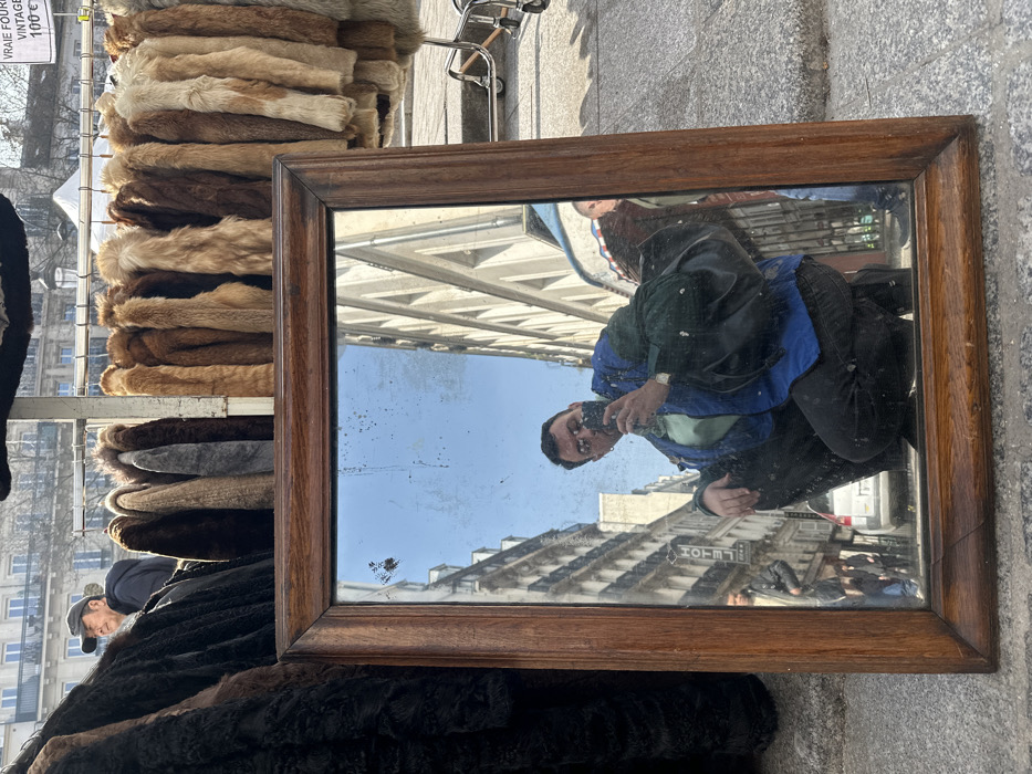 A rectangular mirror with a dark wooden frame rests vertically on a grey stone pavement. The mirror's surface is smudged and shows several small blemishes. Reflected within the mirror is a male individual, partially cropped, in a squatting or crouching position, holding a black smartphone to take a photograph. He wears round glasses, a blue jacket over a light green shirt, dark pants, and a black bag slung over his left shoulder. A watch is visible on his left wrist.
The reflection also reveals a Parisian street scene: multi-story, light-colored Haussmannian buildings with classic window designs and balconies line a thoroughfare. A partially visible street sign for "HOTEL" is discernible on one building. Vehicles and other pedestrians are faintly reflected in the distant background of the mirror. The sky reflected above is clear and bright blue.
Above and behind the mirror, outside the reflection, a metal rack displays numerous vintage fur coats of varying brown, beige, and grey hues. To the left, a section of dark fur coats is visible. A male figure wearing a dark cap and jacket stands behind the fur coats on the left, looking towards the right. To the upper right, a white sign with black text is partially visible, indicating "VRAIE FOURRURE VINTAGE 100€" (True Fur Vintage 100€). The overall scene depicts an outdoor market or flea market (brocante) in Paris, France.