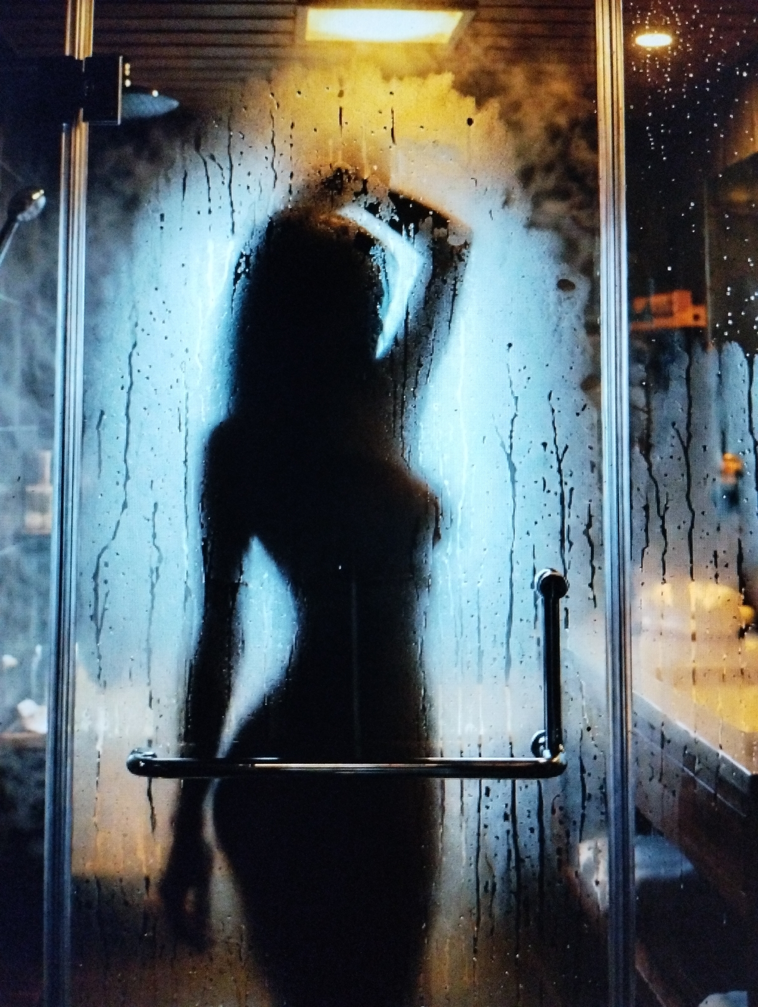 An image shows the silhouette of a person behind a steamy glass shower door. The person's hand is raised and touching their head. Water droplets are visible on the glass, and a blue light illuminates the scene. A metal showerhead is visible on the left side of the image. A chrome handle is attached to the shower door. The background is dark and out of focus, with hints of what appear to be hotel room decor.