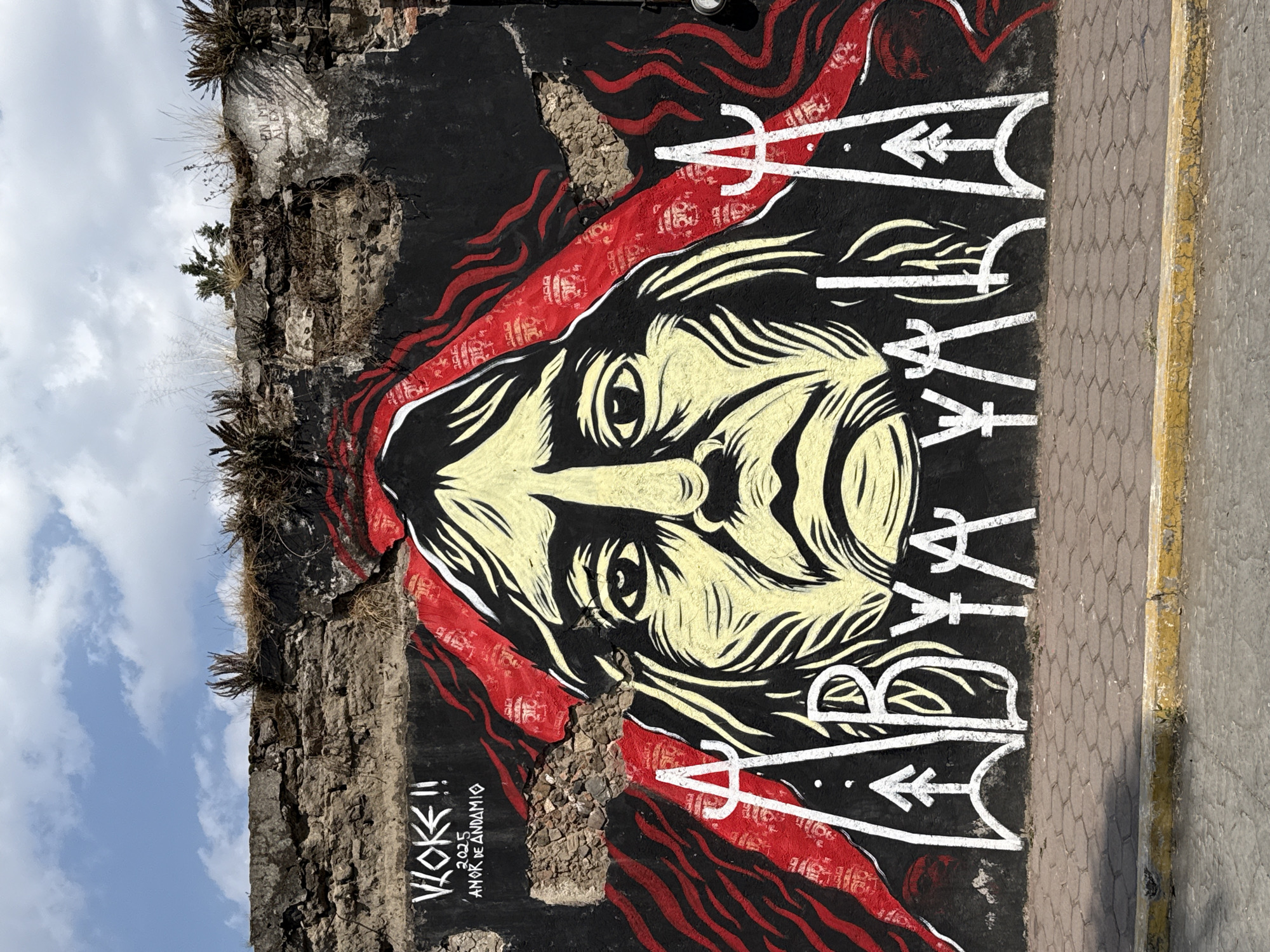 A mural in San Andrés Cholula, Mexico, adorns a rough, stone wall next to a brick-paved surface. The mural depicts a face with stylized, flowing features in black, red, and cream. The face is framed by red patterns resembling hair or fabric. White, geometric shapes and symbols are vertically arranged to the right of the face. Below the mural, near the bottom left corner, are the words "VLOKE!!", the date "2025," and the phrase "AMOR DE ANDAMIO." The stone wall supporting the mural is textured and shows signs of age with small plants growing from its crevices. The sky is visible in the upper left corner.