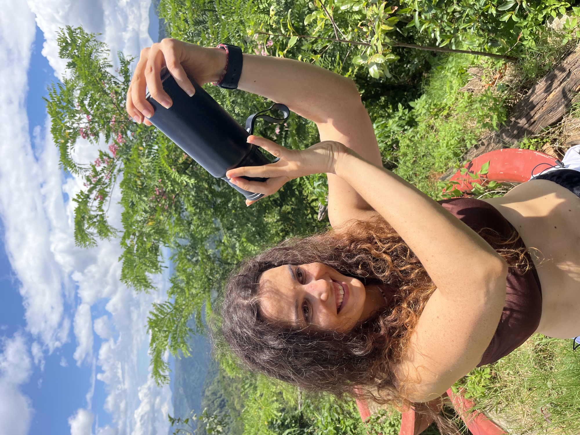 A young woman with curly brown hair and a brown bikini top is taking a selfie with a black water bottle held in her hands. She is outdoors in a lush, green environment with mountains visible in the background under a blue sky with white clouds. The woman is smiling at the camera. The location is identified as Sơn Điền, Vietnam.
