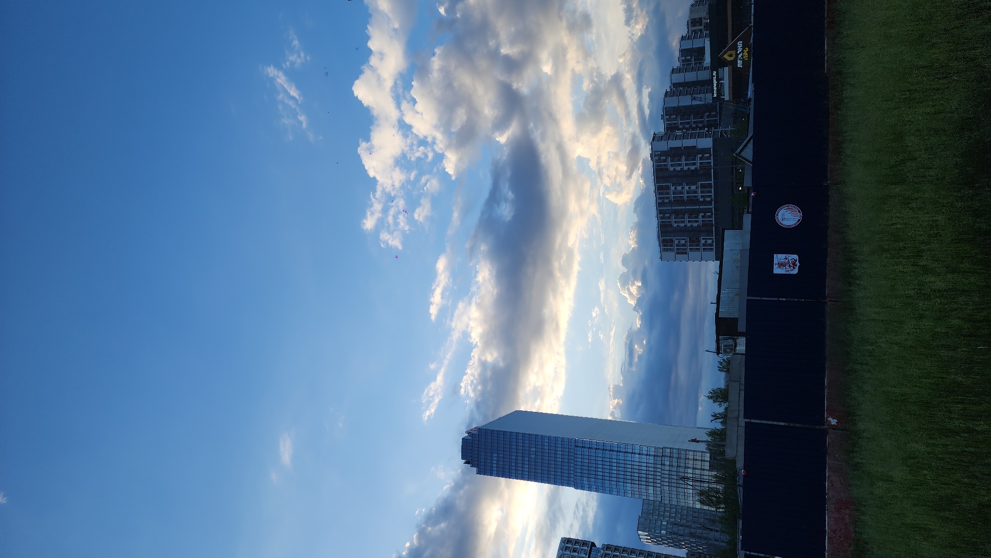 The image shows a view in Moscow, Russia, under a sky filled with clouds. The photograph captures the architectural landscape with a tall, modern glass building as a prominent feature, contrasted by other apartment blocks. A dark fence adorned with sports logos and signage separates the built environment from a green, grassy area. The scene is dominated by the sky and the presence of urban development.