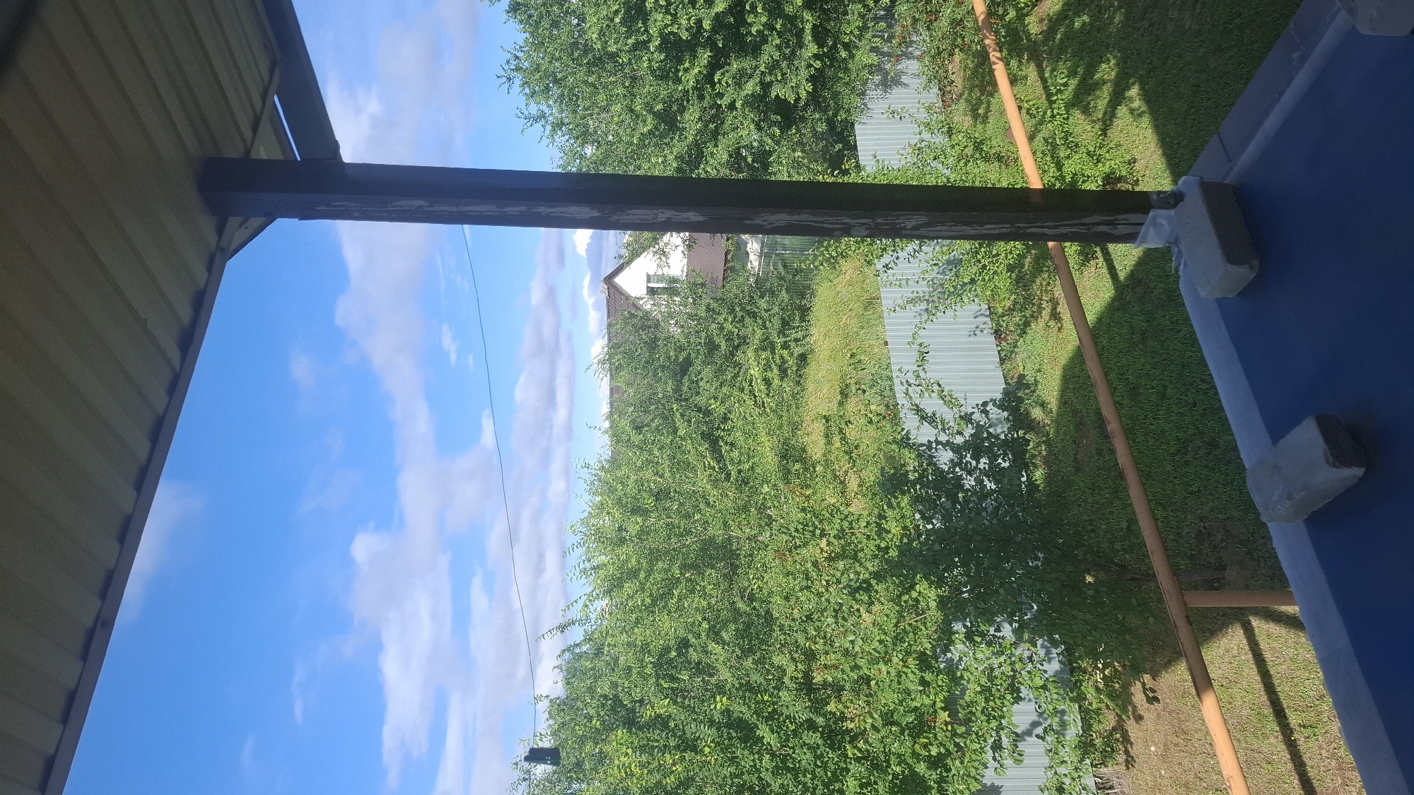 This image captures a view from a covered porch or balcony, looking out onto a residential area on a clear, sunny day. The foreground shows a dark blue surface with two concrete blocks resting on it, possibly used as weights. A rust-colored railing runs horizontally across the mid-ground, casting a shadow. Beyond the railing, a corrugated metal fence encloses a yard filled with lush green trees and vegetation. In the background, a white house with a brown roof is visible through the foliage, suggesting a suburban or rural setting within Engels, Russia. The sky above is a vibrant blue, dotted with scattered white clouds. The overall scene depicts a peaceful, everyday outdoor environment.