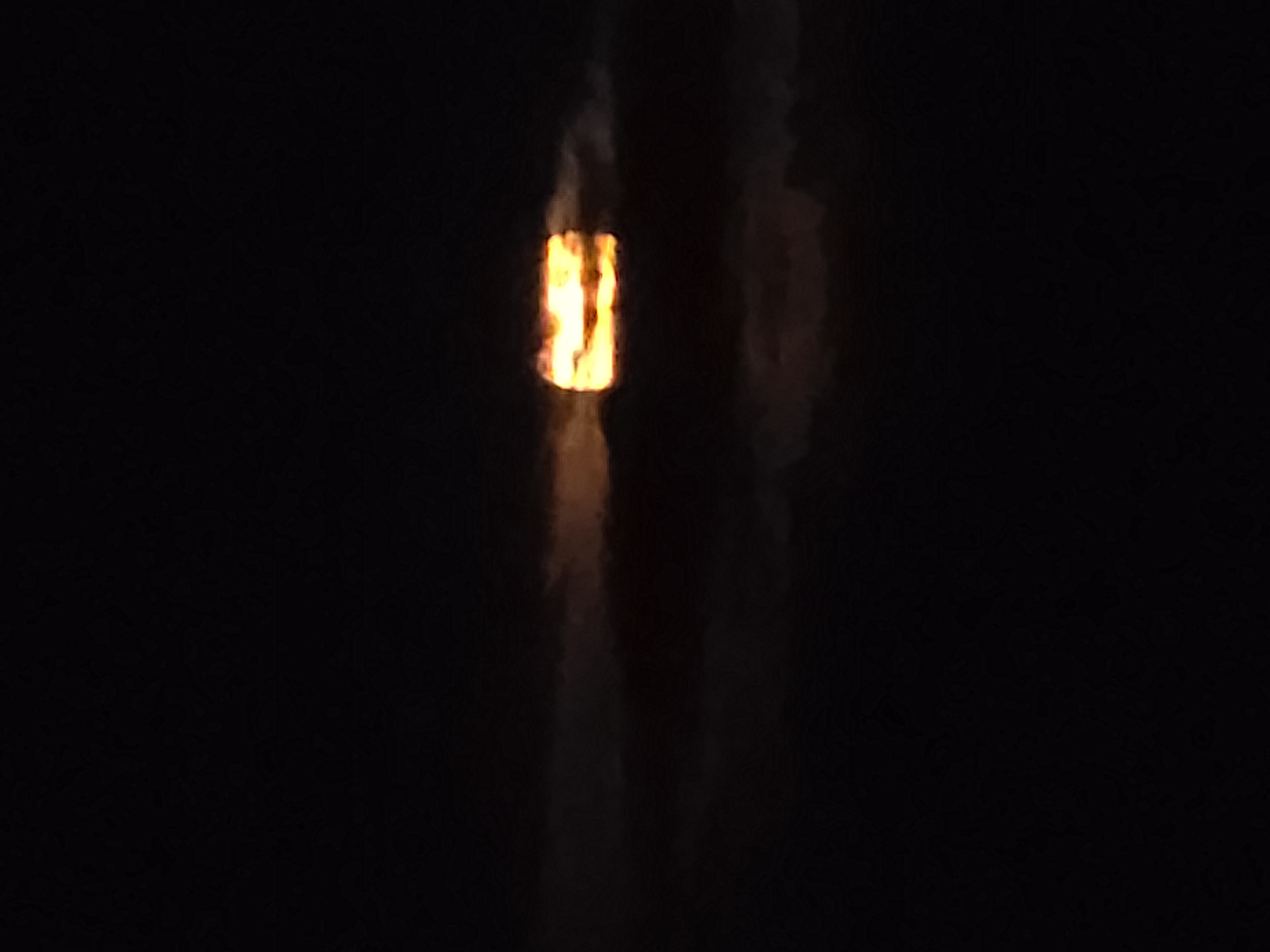 The image captures a highly contrasted scene dominated by a vivid, vertical, flame-like light source against a predominantly dark, almost black background. The central light emanates intense orange and yellow hues, reminiscent of flickering fire or superheated material, and is presented as a distinct rectangular, elongated form. Faint, blurry, dark reddish-brown and gray vertical streaks are subtly visible to the immediate left and right of the bright object, possibly indicating reflections on a nearby surface, smoke, or heat haze. The surrounding environment is characterized by extreme darkness, suggesting either nighttime or an unlit indoor setting. There are no discernible people, specific objects beyond the light source itself, or any visible text. The scene depicts an active emission of light and implied heat within an otherwise obscured location.