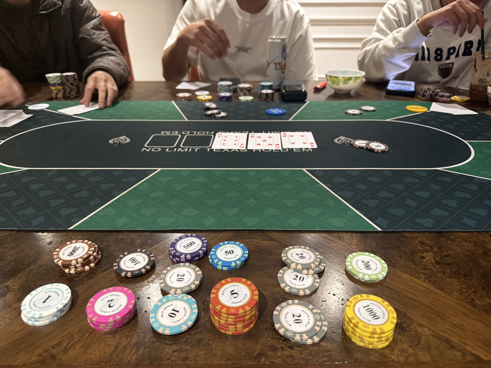 The image depicts a scene of a poker game in progress, set against a wooden table. In the foreground, a collection of poker chips is scattered, with denominations ranging from 1 to 2000. The chips are of various colors and bear the inscription "Monte Carlo Poker Club." One stack of red and yellow chips is labeled with "5," while a stack of white and light blue chips is labeled with "1." A single green chip with a denomination of "200" is also visible.
In the mid-ground, a green poker mat with a diamond pattern is spread out. The mat has a "NO LIMIT TEXAS HOLD'EM" inscription. Several cards are laid out on the mat, including three face-up cards which appear to be a 9 of hearts, a 9 of diamonds, and a 9 of spades, suggesting a potential hand in progress. A small pile of chips is also present on the mat.
In the background, three individuals are partially visible, engaged in the game. The person on the left is wearing a dark jacket and has their hands near some chips and cards. The person in the center, wearing a white t-shirt, is reaching towards the poker mat. The person on the right is wearing a white sweatshirt and is holding a drink. A can of "Tiger" beer and a small white bowl are also on the table. The environment suggests an indoor setting, possibly a home or a casual gaming venue. The image does not provide any specific clues about the location being in Xuân Trường - Đà Lạt, Vietnam.