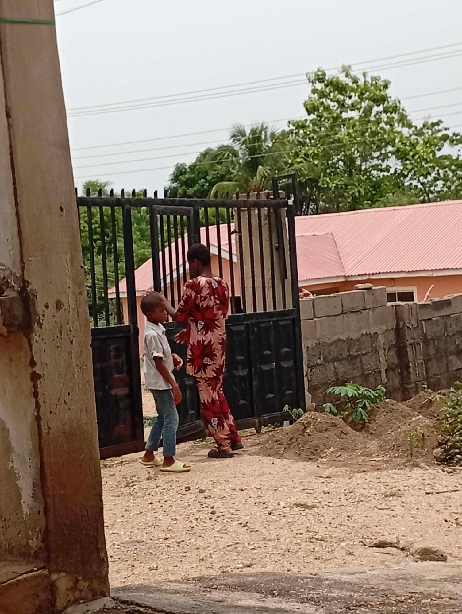 The image depicts two individuals, an adult and a child, standing near a black metal gate in what appears to be a residential area in Adankolo, Nigeria. The child is wearing a light-colored shirt, blue jeans, and yellow sandals. The adult, who is facing the child, is clad in a patterned red and beige ensemble. The gate is set in front of a concrete block wall, with a house featuring a red corrugated roof visible in the background. The ground is covered in gravel. Vegetation is present near the wall and behind the gate, including trees and smaller plants. A concrete structure is partially visible on the left side of the image.