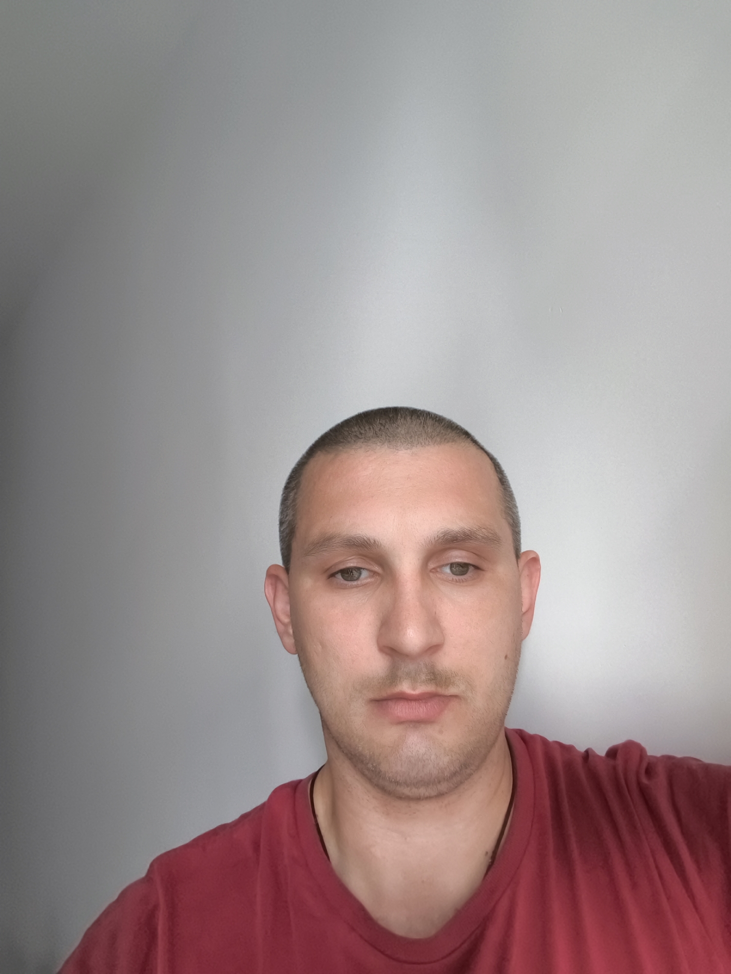 The image is a selfie of a man with short hair, wearing a red t-shirt. He has fair skin and light-colored eyes. The background is a plain, light-colored wall. The image does not contain any recognizable landmarks or contextual information that would allow for identification of a specific location in Wolgast, Germany.