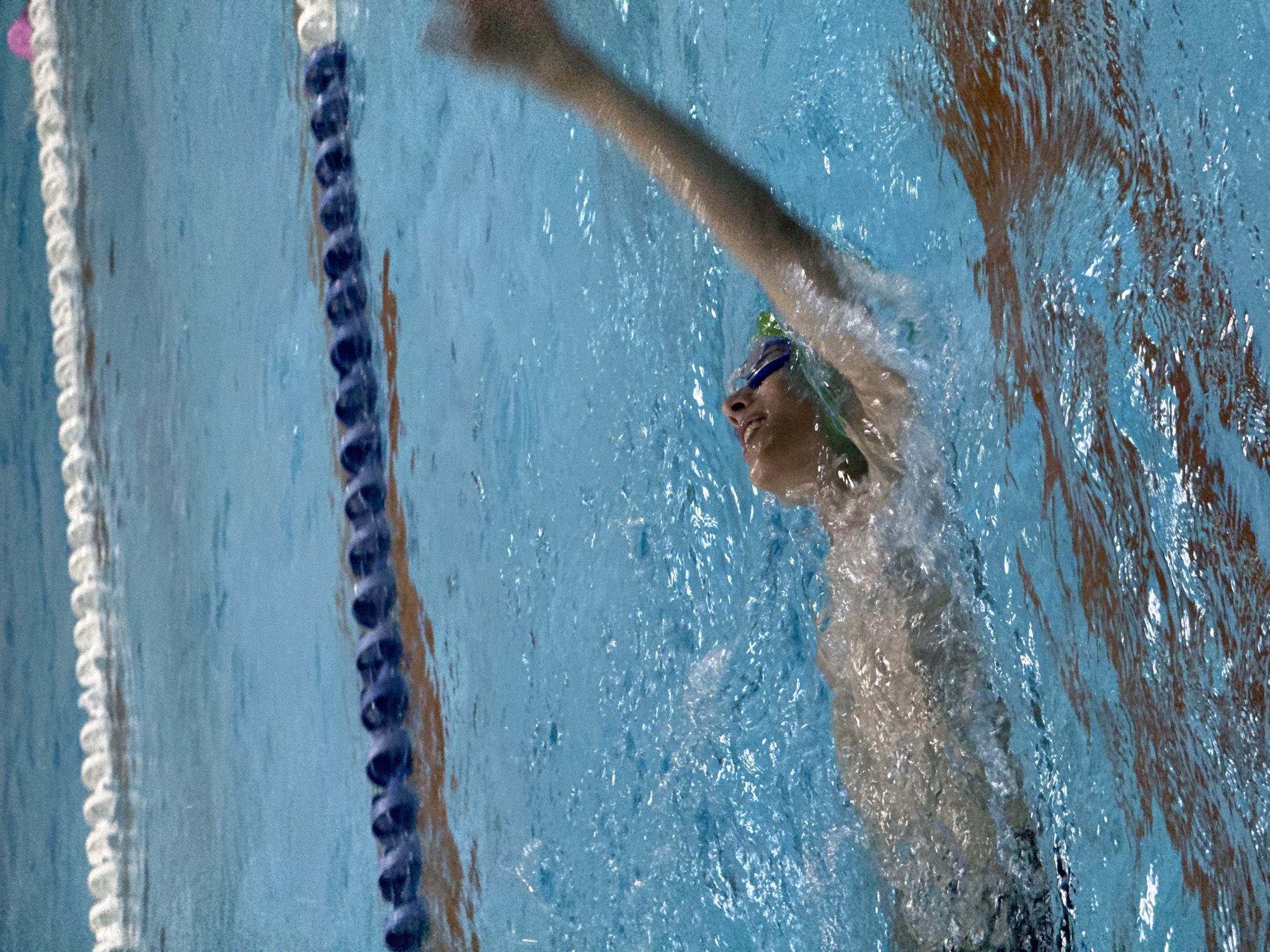 A young person swims on their back in a bright blue pool with lane markers. The person is wearing blue goggles and a green cap. Their right arm is extended out of the water and blurred due to motion. The pool water is disturbed, showing ripples and splashes. The background is out of focus, with a white lane marker at the top and a blue and white lane marker below it. This image was taken in Birobidzhan, Russia.