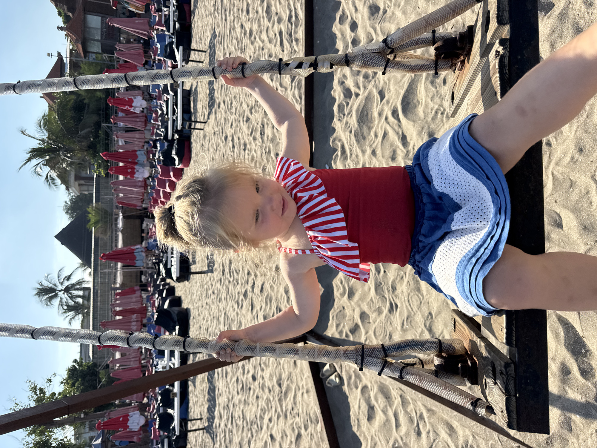 A young blonde child is captured playing on beachside equipment under bright sunlight. The child, likely a girl, is wearing a red top with red and white striped ruffles on the shoulders and blue shorts with white crochet detailing. She is holding onto thick ropes, possibly part of a swing or play structure, which are covered in a clear mesh material and secured with black zip ties. Her expression is neutral, and she is looking towards the viewer.
The setting is a sandy beach, with distinctive ripples and shadows visible in the sand, indicative of strong sunshine. In the background, numerous beach loungers and umbrellas, primarily red with some blue, are neatly arranged in rows. Further behind, lush green palm trees stand against a clear blue sky, with buildings visible in the distance. The overall scene suggests a resort or beach club environment. No discernible text is visible in the image. The time of day appears to be late morning or afternoon, given the bright illumination and shadow patterns. The location context indicates Kuta, Indonesia.