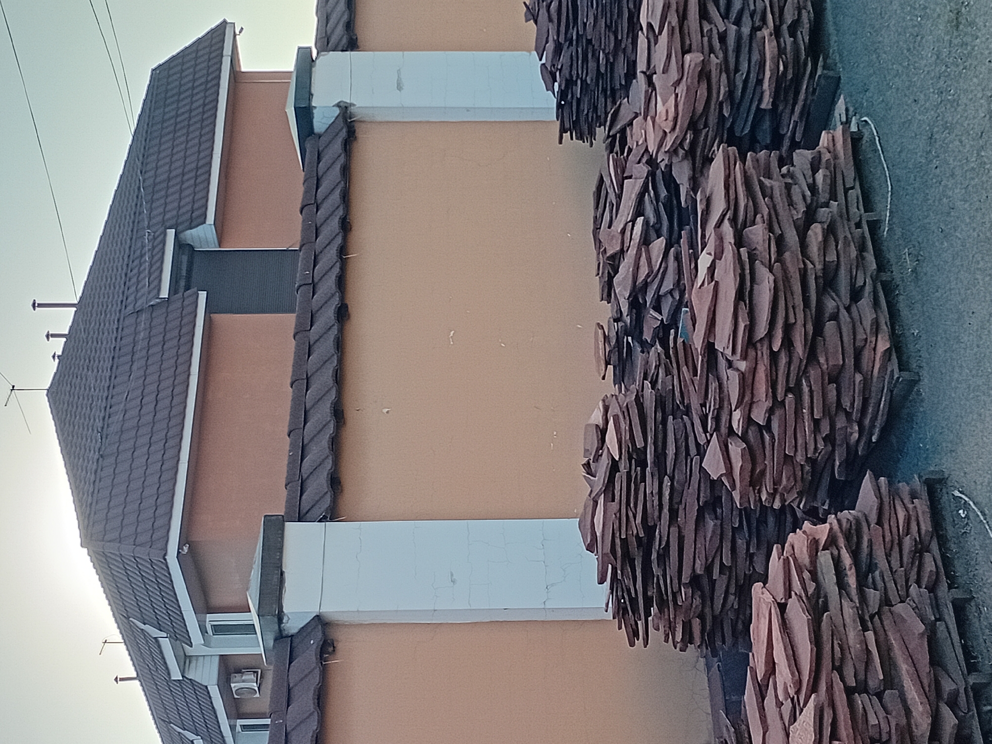The image depicts an outdoor scene in the city of Semey, Kazakhstan, featuring a tall, tan-colored wall with white support columns. The wall is topped with a dark brown, tiled roof that extends to cover a residential building visible behind it. The building has a similarly tiled roof and an external air conditioning unit. In front of the wall, several stacks of flat, reddish-brown stones are piled on wooden pallets. The ground is paved with asphalt. The sky above is a pale, hazy blue, suggesting either early morning or late afternoon light. There are no people visible in the image.