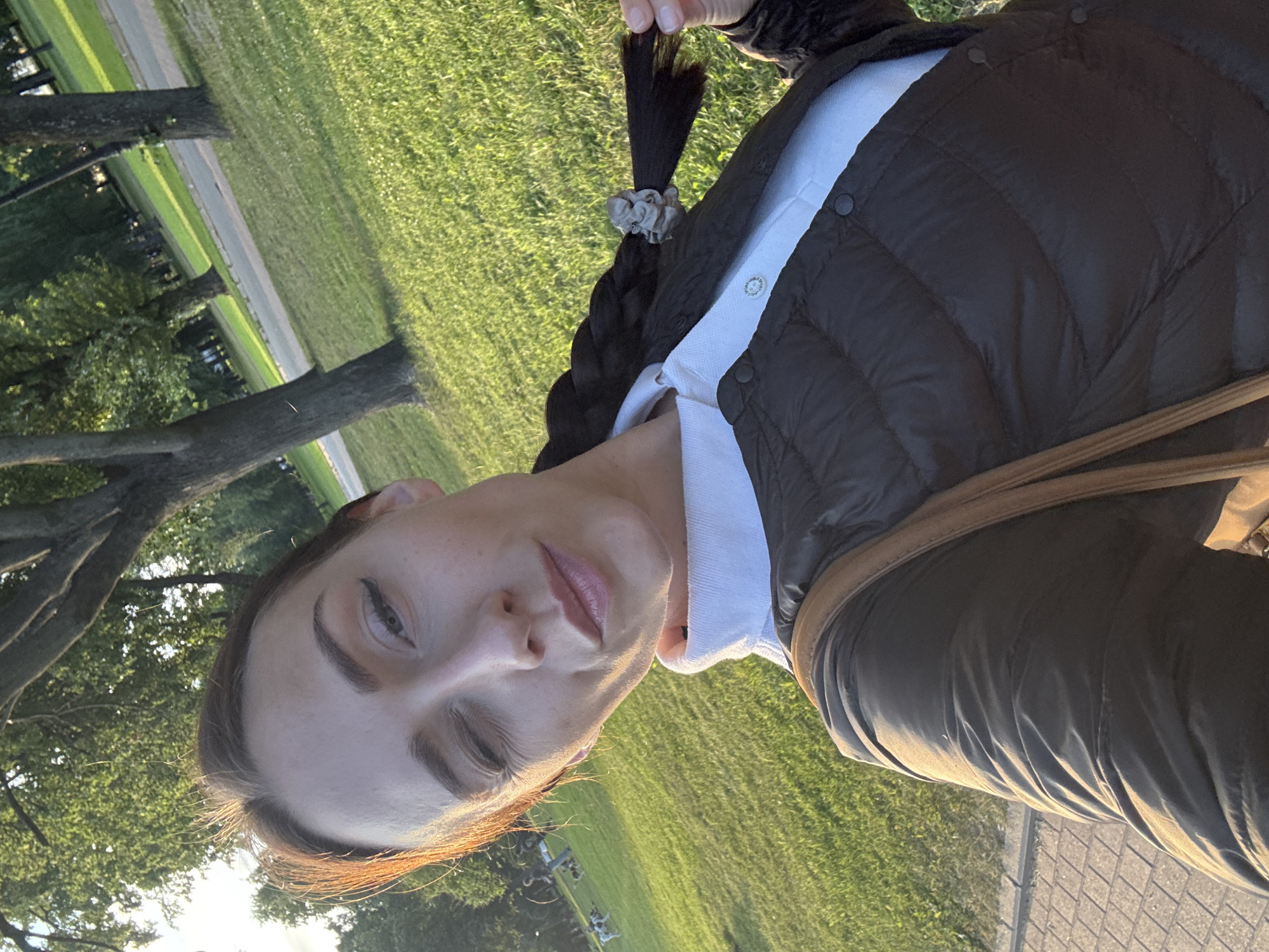 This is a portrait of a woman outdoors, with the image rotated 90 degrees counter-clockwise. The woman is wearing a dark, quilted jacket over a white collared shirt. She has her hair in a braid, secured with a scrunchie. She is looking at the camera with a neutral expression. The background shows a grassy area with trees, suggesting a park or outdoor recreational space. The lighting indicates it is late afternoon or early evening, with the sun casting long shadows and a warm glow. There are no visible text elements in the image. The woman's makeup is subtle, with eyeliner and mascara defining her eyes. The overall scene is casual and relaxed.