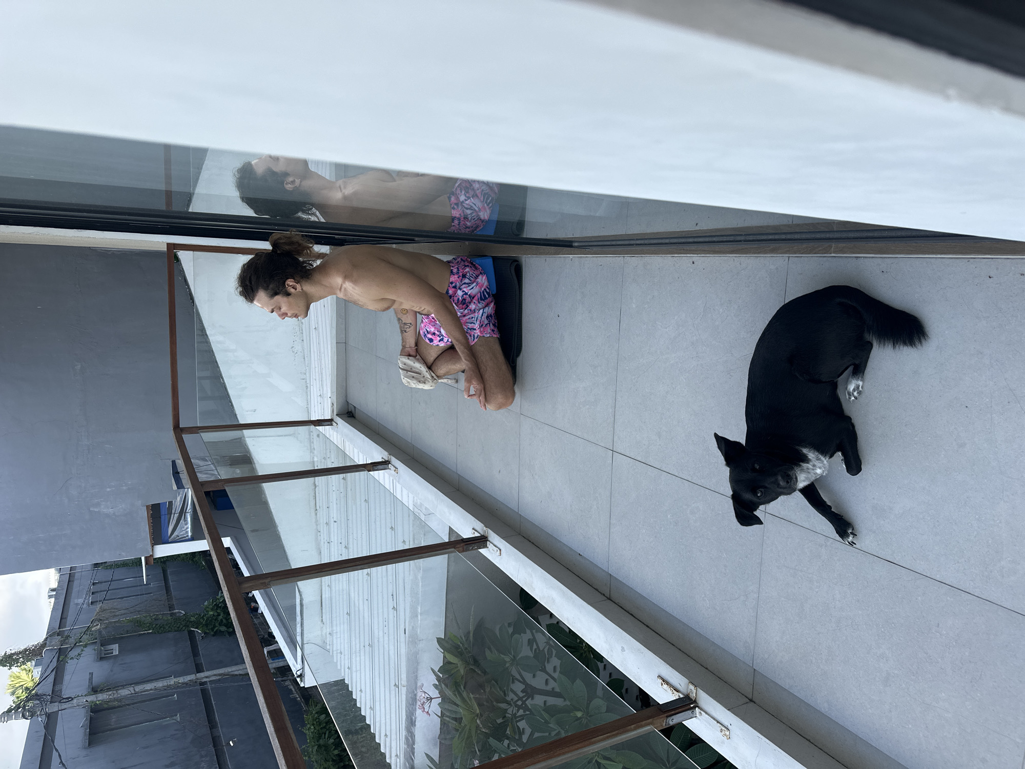 On a balcony in Mengwi, Indonesia, a man with a ponytail is kneeling on a yoga mat in colorful shorts. He is reflected in the window behind him. A black dog with white paws is lying on the tiled floor near the balcony railing. Outside the balcony are trees and a building.
