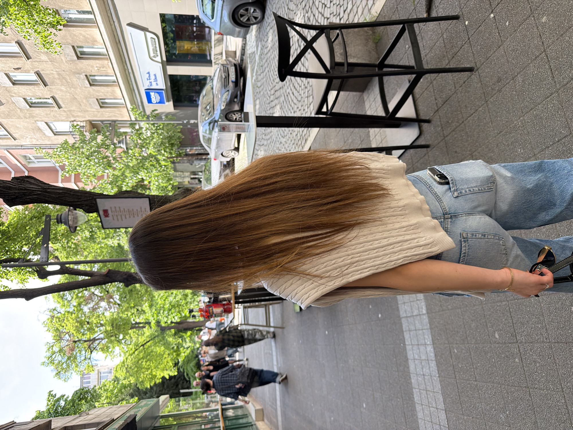 The image depicts a person with long brown hair standing on a sidewalk in Sofia, Bulgaria. They are wearing a light-colored, textured sweater and blue jeans with a pocket visible in the back. A pair of sunglasses is held in their right hand, and a gold bracelet is visible on their wrist. The sidewalk is paved with gray rectangular stones. In the background, there is outdoor seating with black metal chairs and tables, with people seated and standing. Cars are parked on the street, and buildings with multiple windows are visible on the left side of the frame. Trees with green leaves line the street, and a street sign or menu board is attached to one of the trees. The scene appears to be a busy street with outdoor cafes and shops in Sofia, Bulgaria.