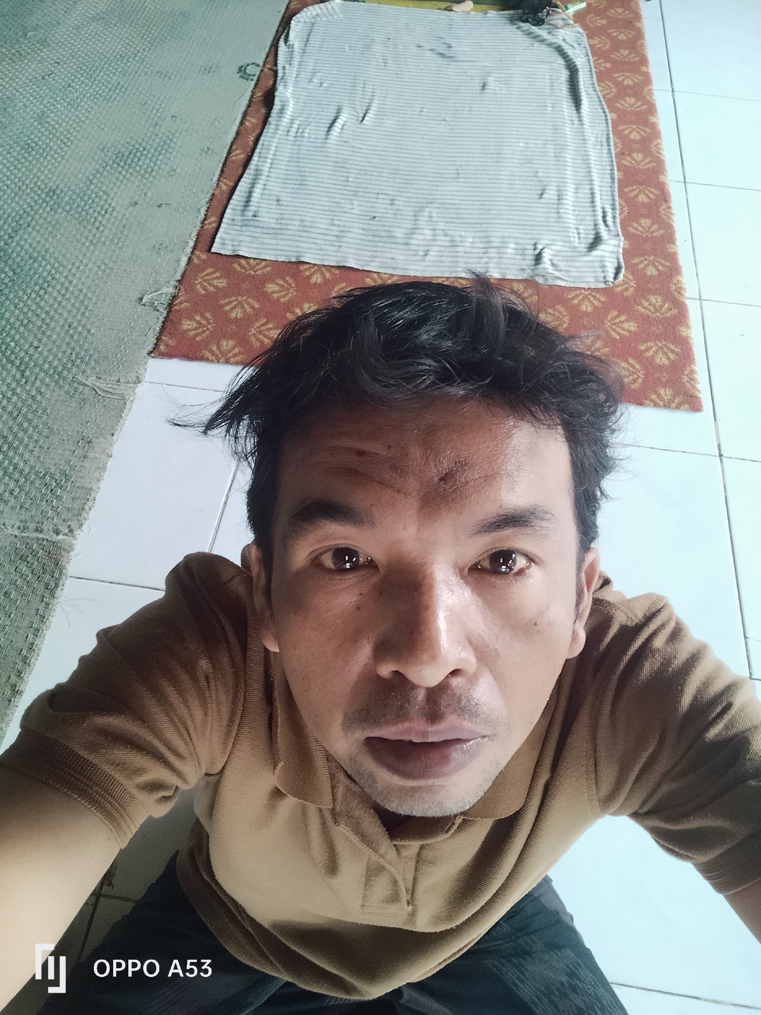 A medium shot selfie shows a man with dark, unkempt hair and brown skin looking directly at the camera. He is wearing a brown polo shirt and dark pants. His mouth is slightly open. In the background, there is a striped t-shirt on a patterned rug, and a greenish textured mat on the left. The floor appears to be tiled. The scene is indoors. The device used to capture the image is an "OPPO A53". The image was taken in Badung, Indonesia.
