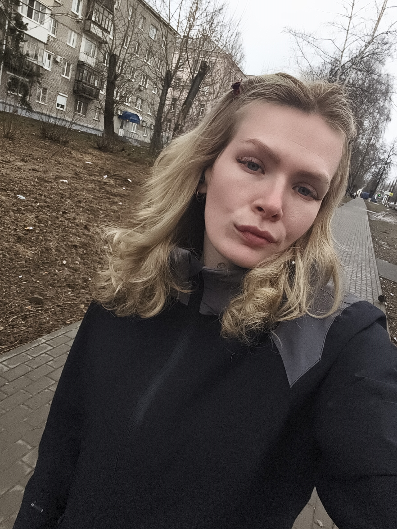 A young, light-skinned woman with blonde, wavy hair is taking a selfie outdoors. She is wearing a dark jacket with grey accents on the shoulders. She has fair skin, slightly defined eyebrows, and a subtle lip color. The background shows a typical urban environment. To the left is a grassy area with what appears to be dead leaves and some trash. Behind that is a multi-story apartment building constructed of grey brick. Several bare trees are visible in front of the building. To the right, there's a paved sidewalk with a pattern of rectangular bricks. Beyond the sidewalk, additional bare trees line the street. The overall scene suggests it is a cloudy day, possibly early spring or late autumn. Based on the style of architecture in the background this scene is located in the city of Izhevsk, Russia.