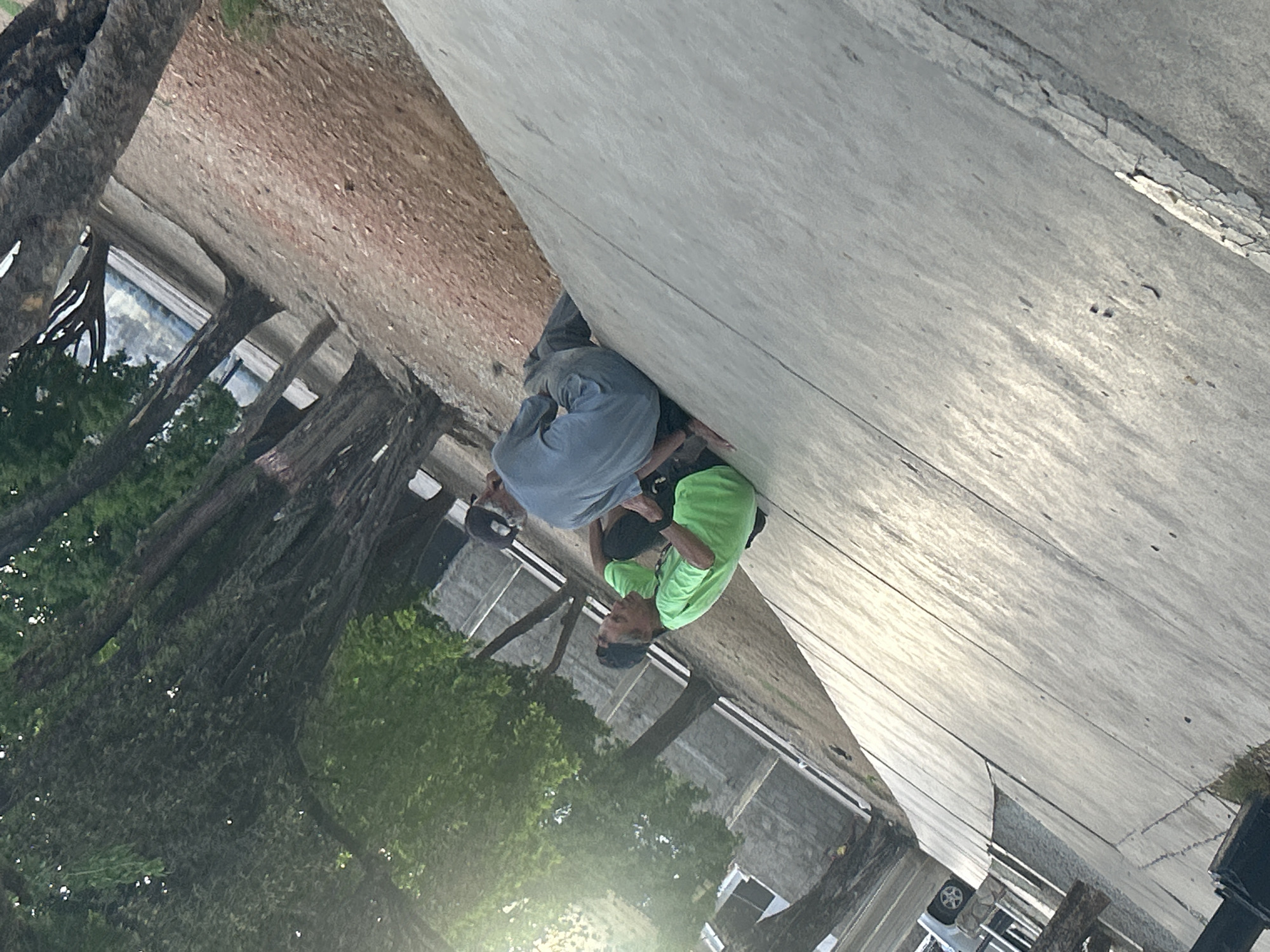 Two individuals are seated on a concrete surface near a dirt path, with trees lining the background. One person, wearing a bright green t-shirt and dark pants, is looking upwards and to the left. The other individual, dressed in a blue shirt and dark pants, is facing away from the camera, leaning back slightly. The scene appears to be outdoors, possibly in a park or a public area. The lighting suggests it is daytime, with sunlight filtering through the trees. The overall impression is of a casual, relaxed moment. The location in the city of Santa Ana, Venezuela is not discernible from the image itself.