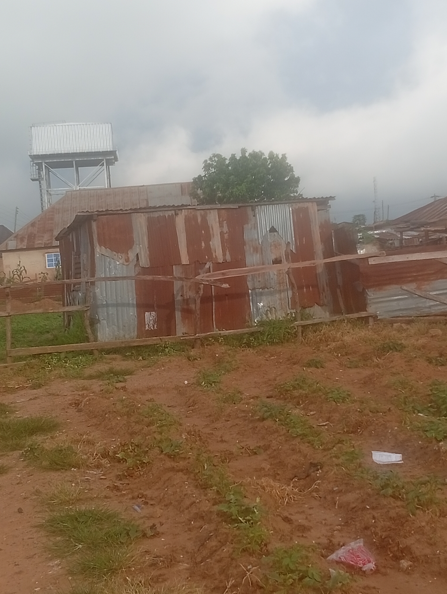 This image depicts an outdoor scene in Bokkos, Nigeria, characterized by a slightly overcast sky, suggesting it might be morning or late afternoon. The environment appears somewhat arid with reddish-brown soil and patches of dry and green grass.
The most prominent structures are makeshift buildings constructed from corrugated metal sheets. These structures are weathered and rusty, indicating they are likely older or exposed to the elements. One building in the foreground is particularly striking with its patchwork of different metal sheets and a leaning wooden beam across the top. Behind these, a more substantial building with a pitched, rusty metal roof is visible, and further back, a large, modern-looking water tower stands tall against the sky. This water tower has a metallic, ribbed tank supported by a metal frame.
The scene seems to be in a rural or semi-urban area, possibly a residential or commercial area with informal structures. There is no visible human activity or clear event taking place. The overall impression is one of a simple, perhaps less developed, settlement. A few pieces of litter are scattered on the ground in the foreground. One white piece of paper or plastic is visible, and another is a plastic bag containing what appears to be red meat. There is no visible text in the image.