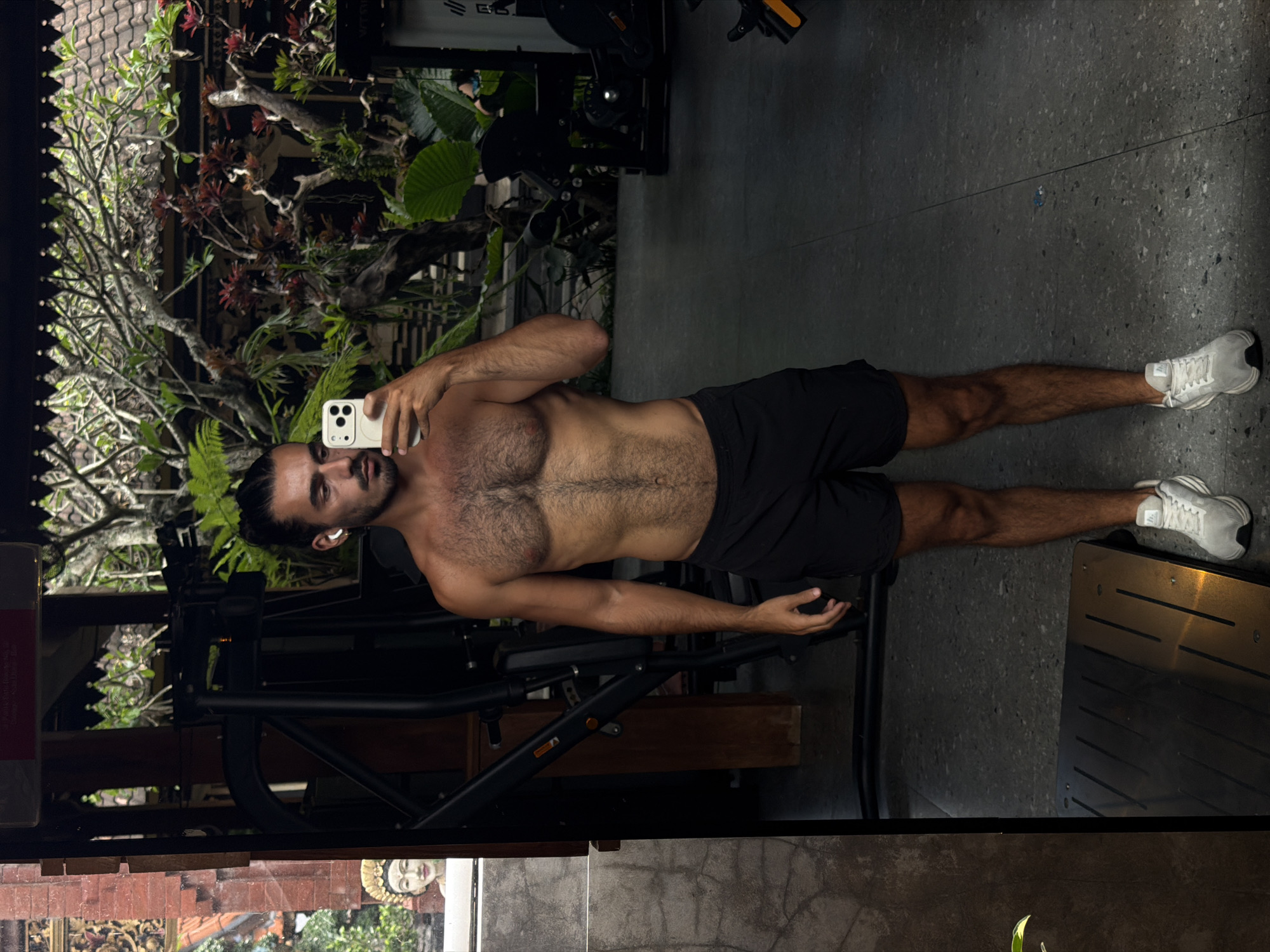 A man stands in what appears to be a gym, taking a selfie. He is shirtless, wearing black shorts, and white sneakers. He holds a white smartphone in his left hand. He has dark hair, a beard, and is wearing a white earbud in his left ear. Behind him is gym equipment and a view of lush greenery, including a tree and various plants. The gym appears to be connected to an outdoor area. The reflection in the mirror shows an ornamental carved image. This scene appears to be in North Kuta, Indonesia, given the tropical setting and Balinese cultural elements in the decor.