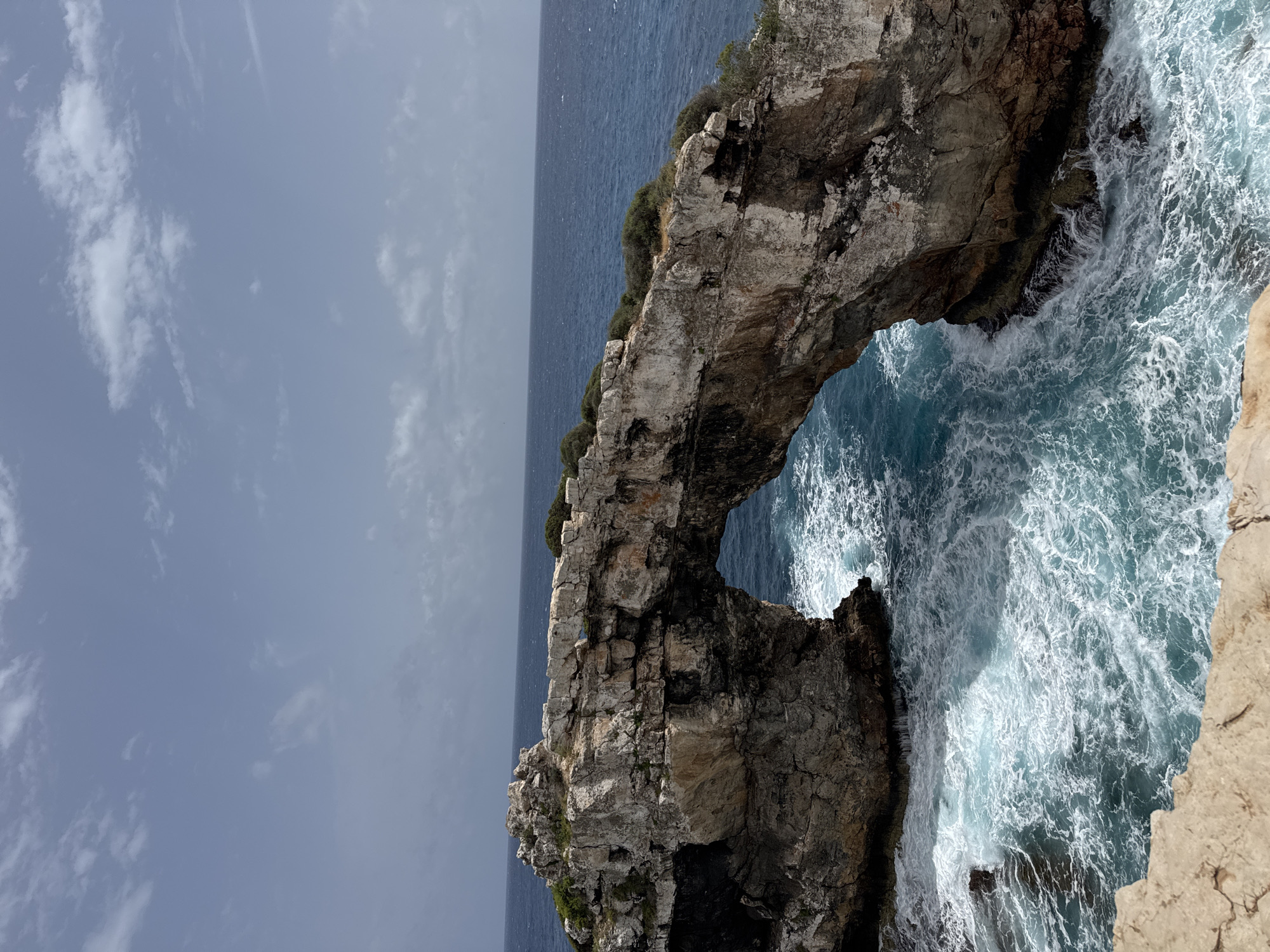 The image shows Es Pontàs, a natural rock arch located off the coast of Santanyí, Spain. The arch, made of light grey and tan rock, extends over the turquoise and white-capped ocean. A narrow band of vegetation grows on top of the arch. The sky is light grey with some small clouds. Waves crash against the rock formations. The image is oriented vertically.