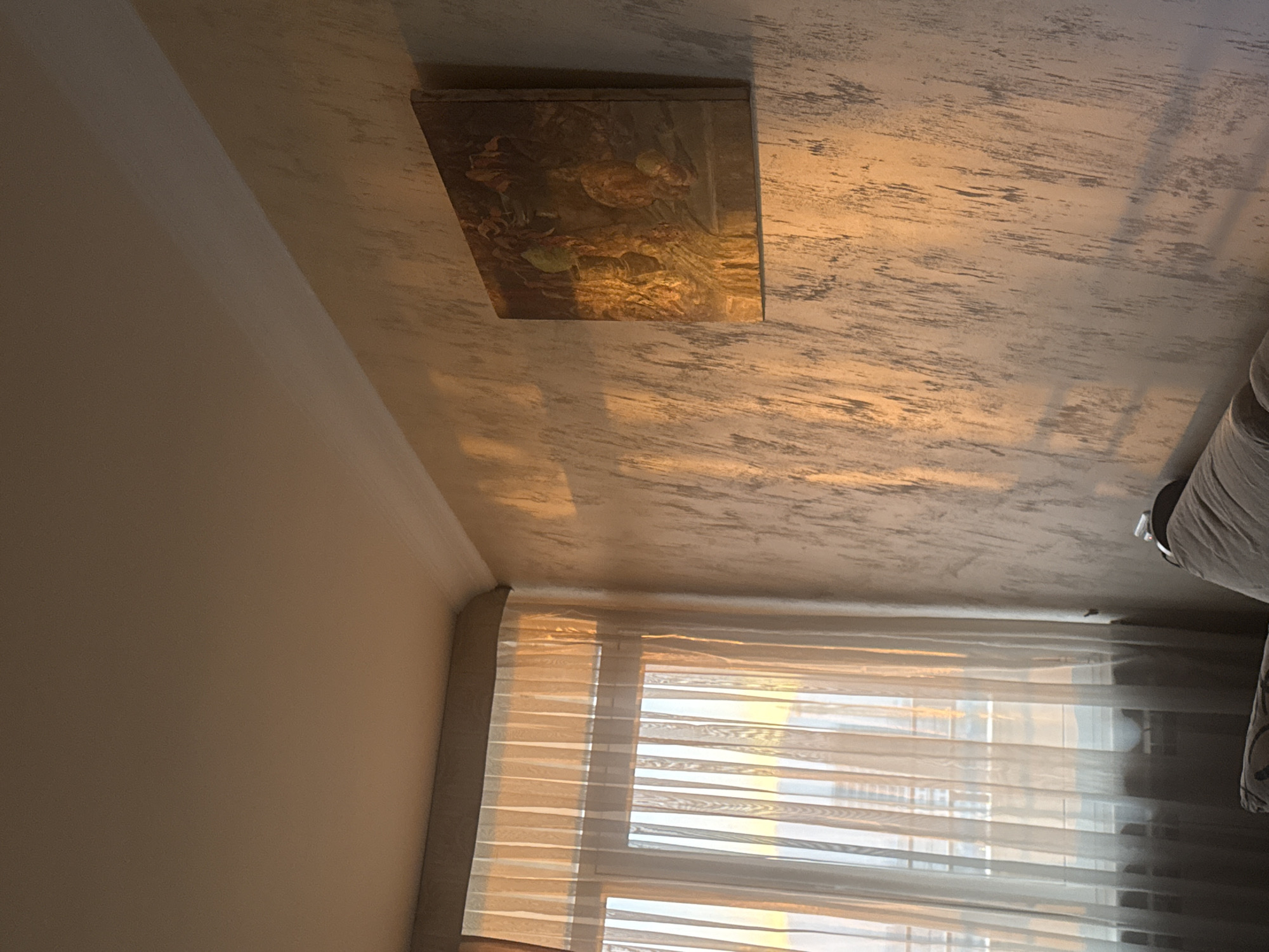 A low-angle indoor shot shows a room in Abovyan, Armenia, with a textured ceiling, a rectangular framed artwork, a wall, a window covered with translucent blinds, and a partial view of a couch. The light source is likely natural sunlight coming through the window, creating shadows and highlights throughout the room. The color palette consists of warm tones of beige, brown, and white. The composition is static, with the focus on the architectural details and interior elements.