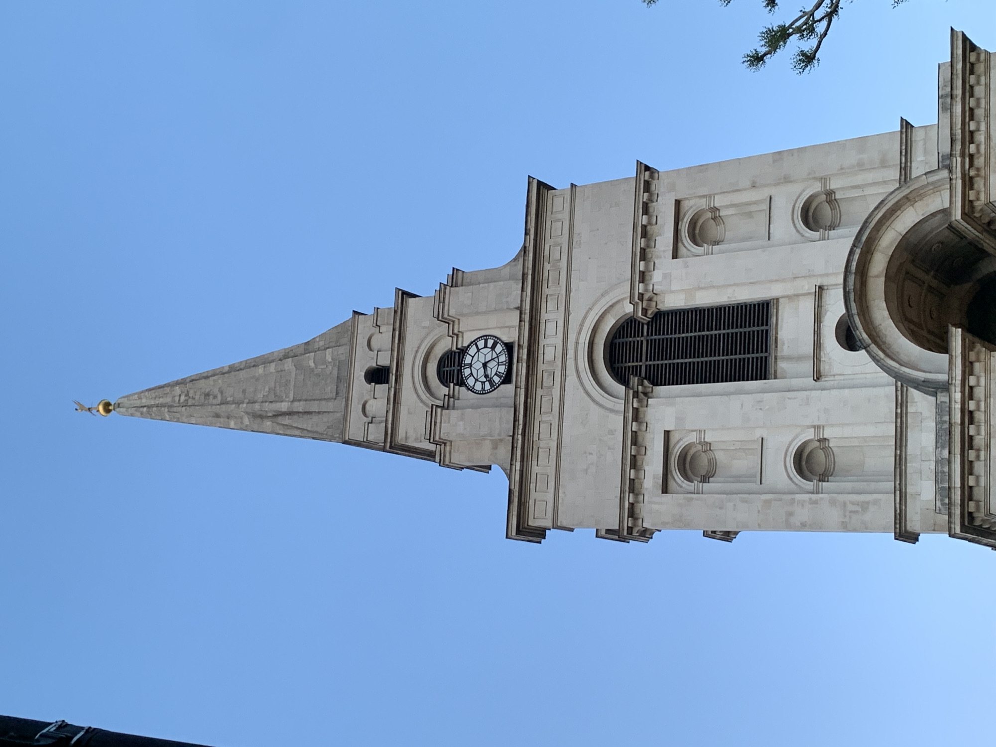 A low-angle shot captures the ornate stone spire and upper sections of a prominent building, likely a church or clock tower, set against a cloudless, bright blue sky. The structure, made of light-colored stone, exhibits classical architectural features including decorative pilasters, cornices, and recessed circular and rectangular elements. A clock face, with clear Roman numerals and hands, is visible on one side of the tower, indicating a time around 11:55 or 12:55.
The tower tapers into a tall, pointed spire, crowned with a small, golden finial or weather vane at its apex. A few green tree branches are visible in the upper right quadrant of the image. The clear sky and strong lighting suggest a sunny daytime setting. The architecture is consistent with historical buildings found in London, United Kingdom, where this image was taken, specifically resembling the spire of St Mary-le-Bow church. No discernible activity is taking place, and there is no visible text.